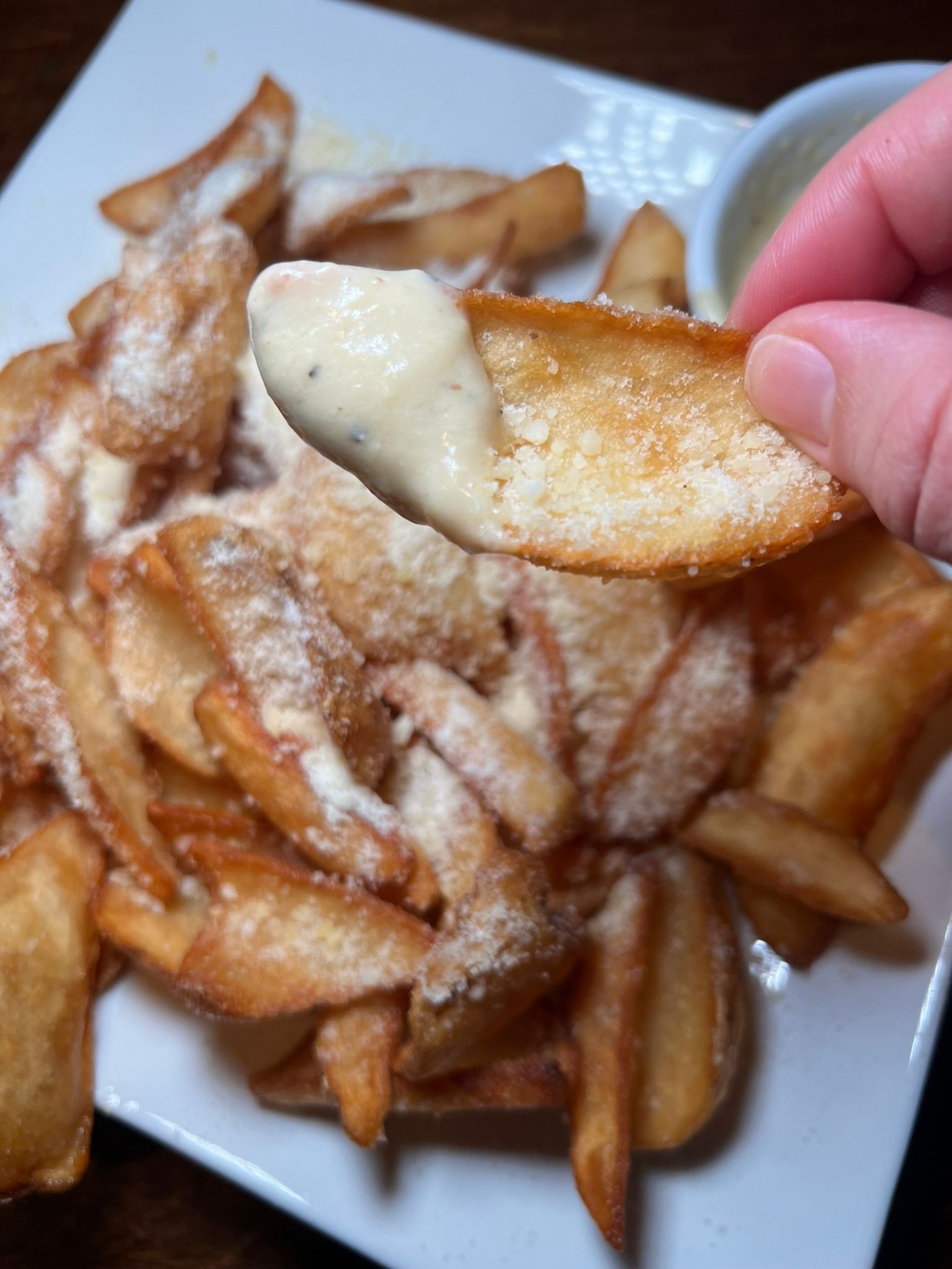 Hand holding a sugar-dusted potato wedge over a plate of fried wedges with dipping sauce