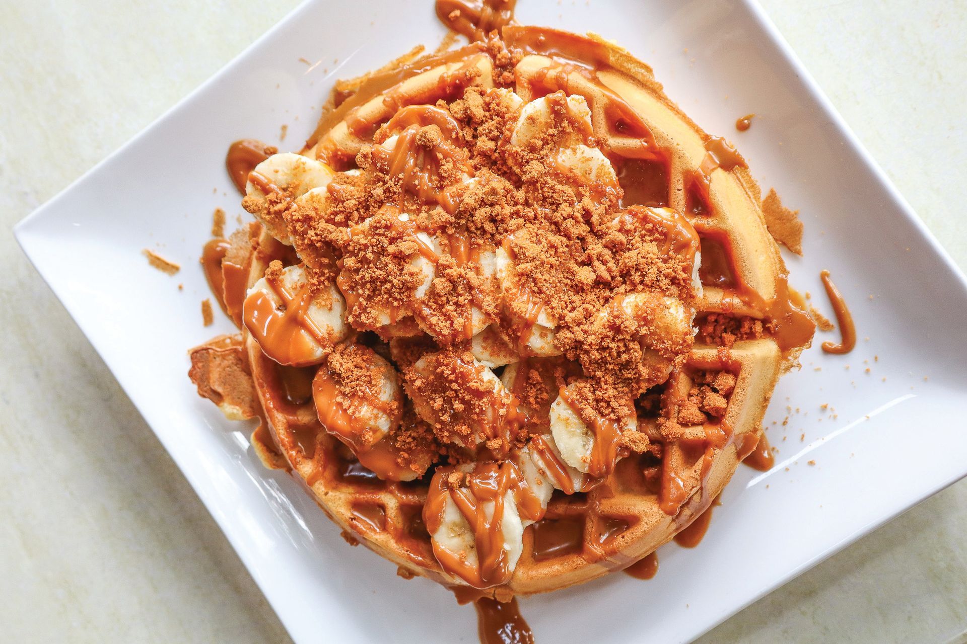 Waffle topped with banana slices and crumbly sauce on a white square plate