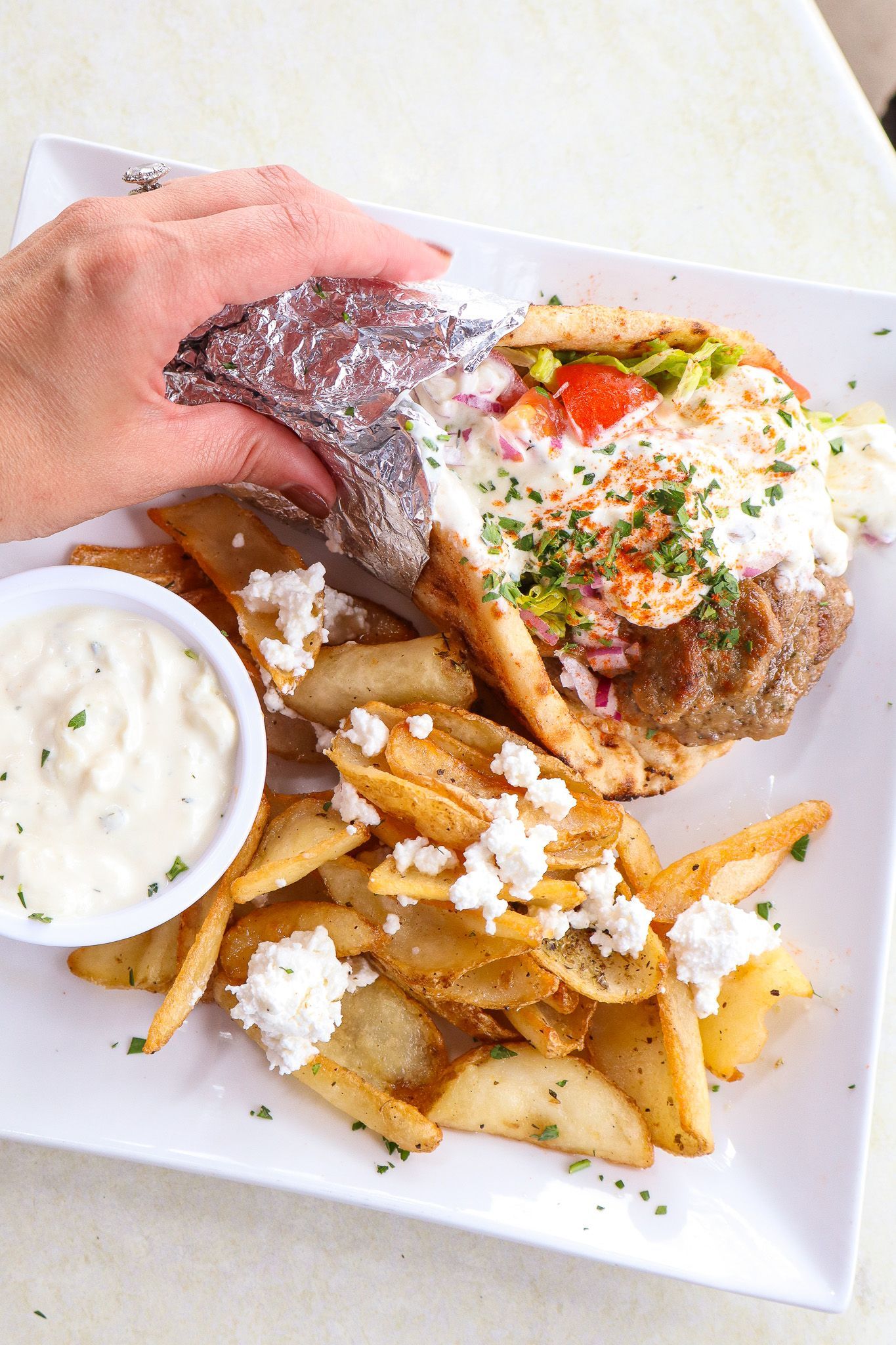 Hand holding a lobster roll with fries and a cup of creamy dipping sauce on a white plate