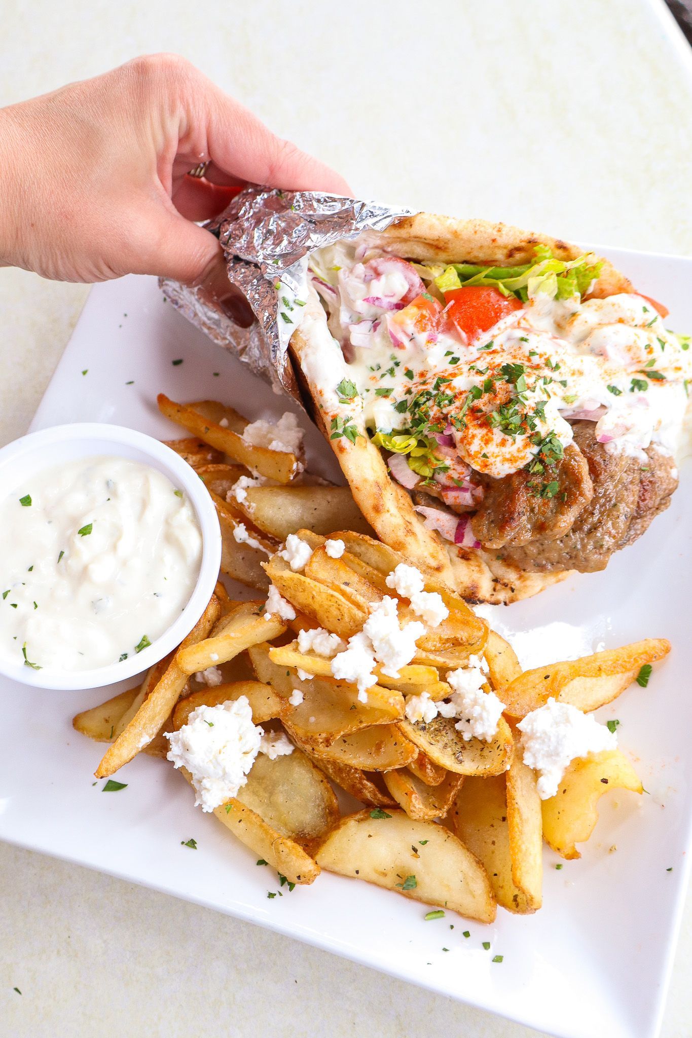 Hand holding a loaded sandwich with fries and dipping sauce on a white plate