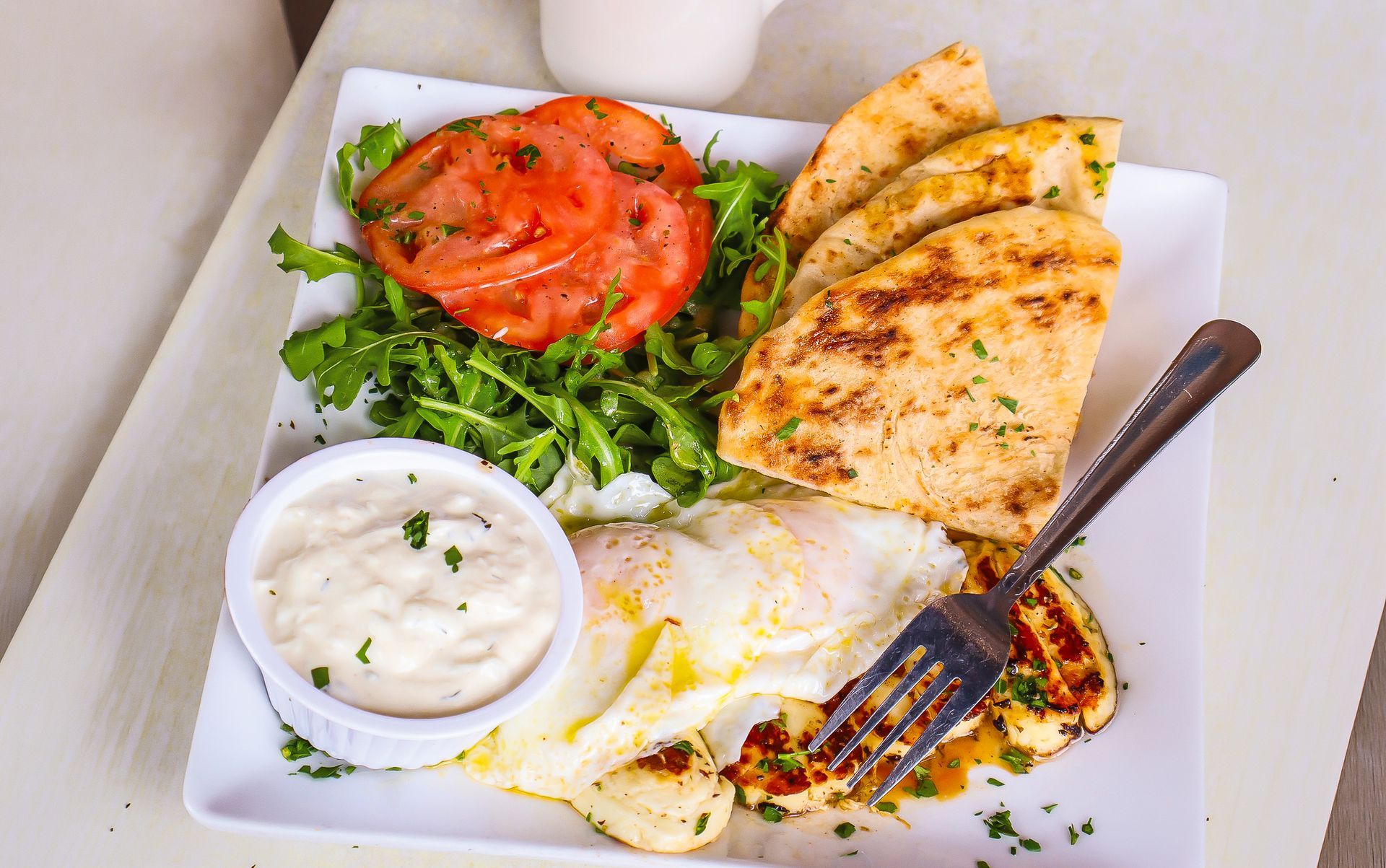 Plate of Mediterranean appetizer with hummus, fried eggs, tomato, greens, pita bread, and a fork
