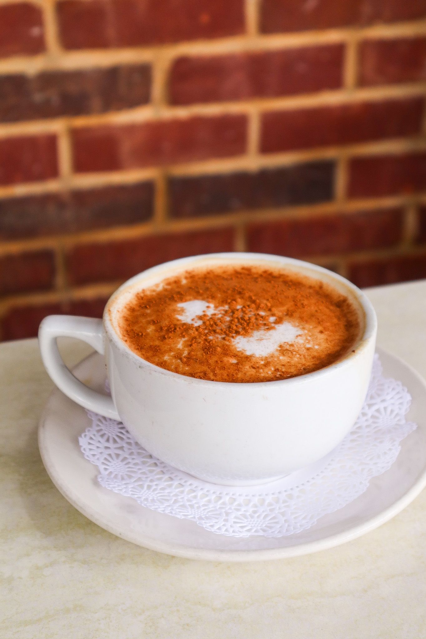White cup of cappuccino with foam on a saucer, set on a table against a brick wall.