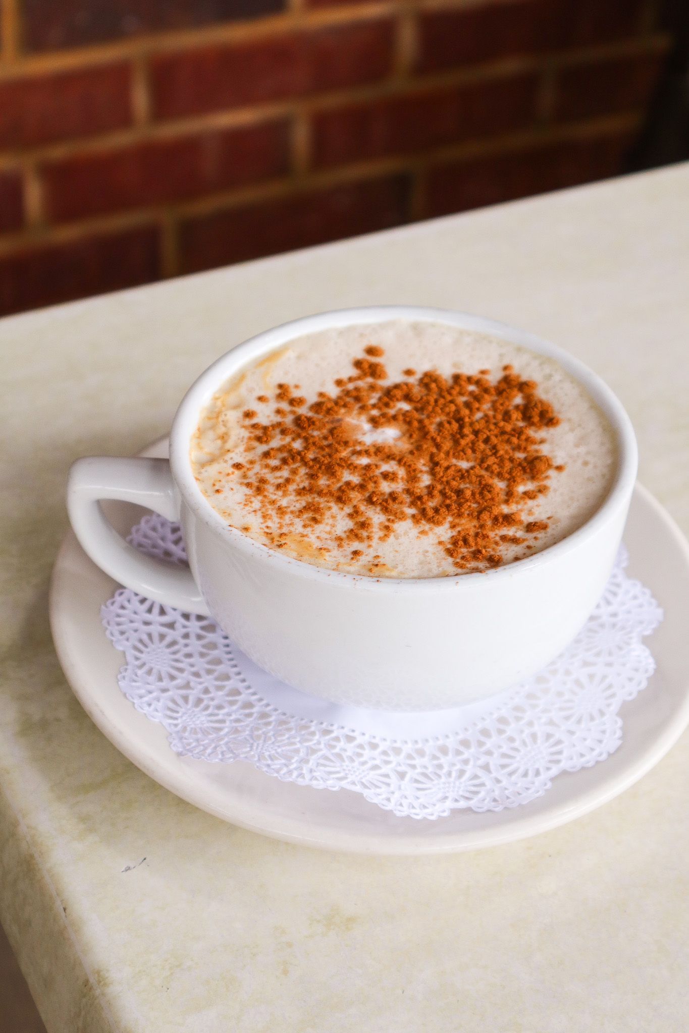 White coffee cup with frothy drink and cinnamon on a saucer atop a table, against a brick wall.
