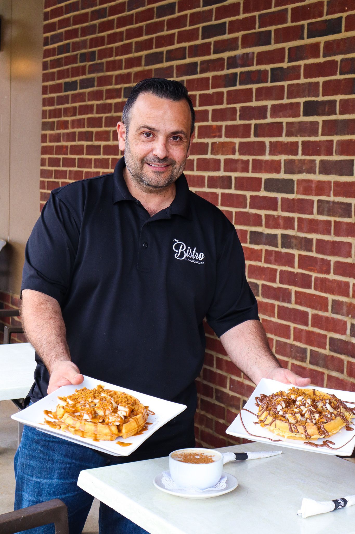 Man holding two plates of food beside a table with coffee, standing against a brick wall