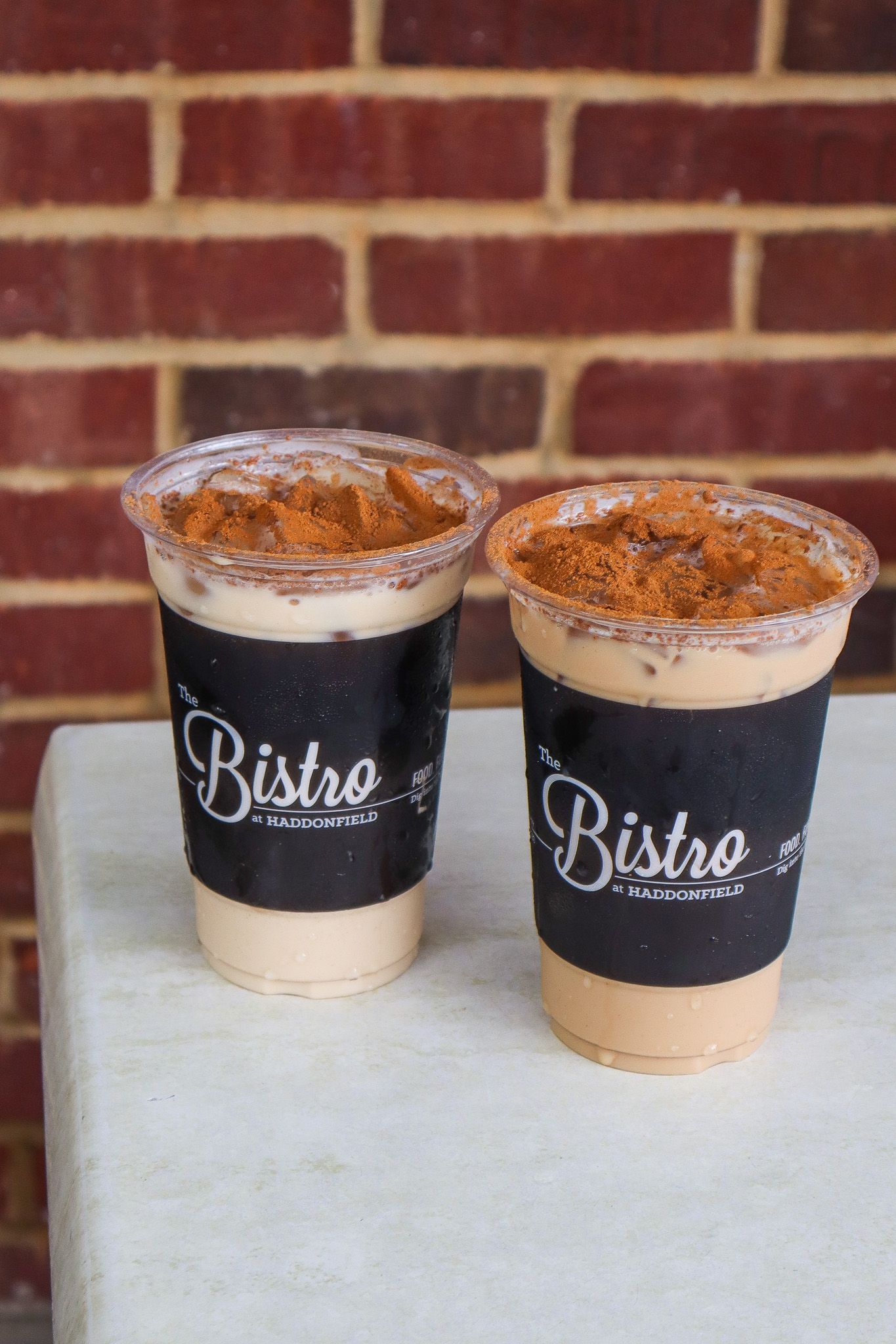 Two Bisko-branded iced drinks in plastic cups on a table against a brick wall.