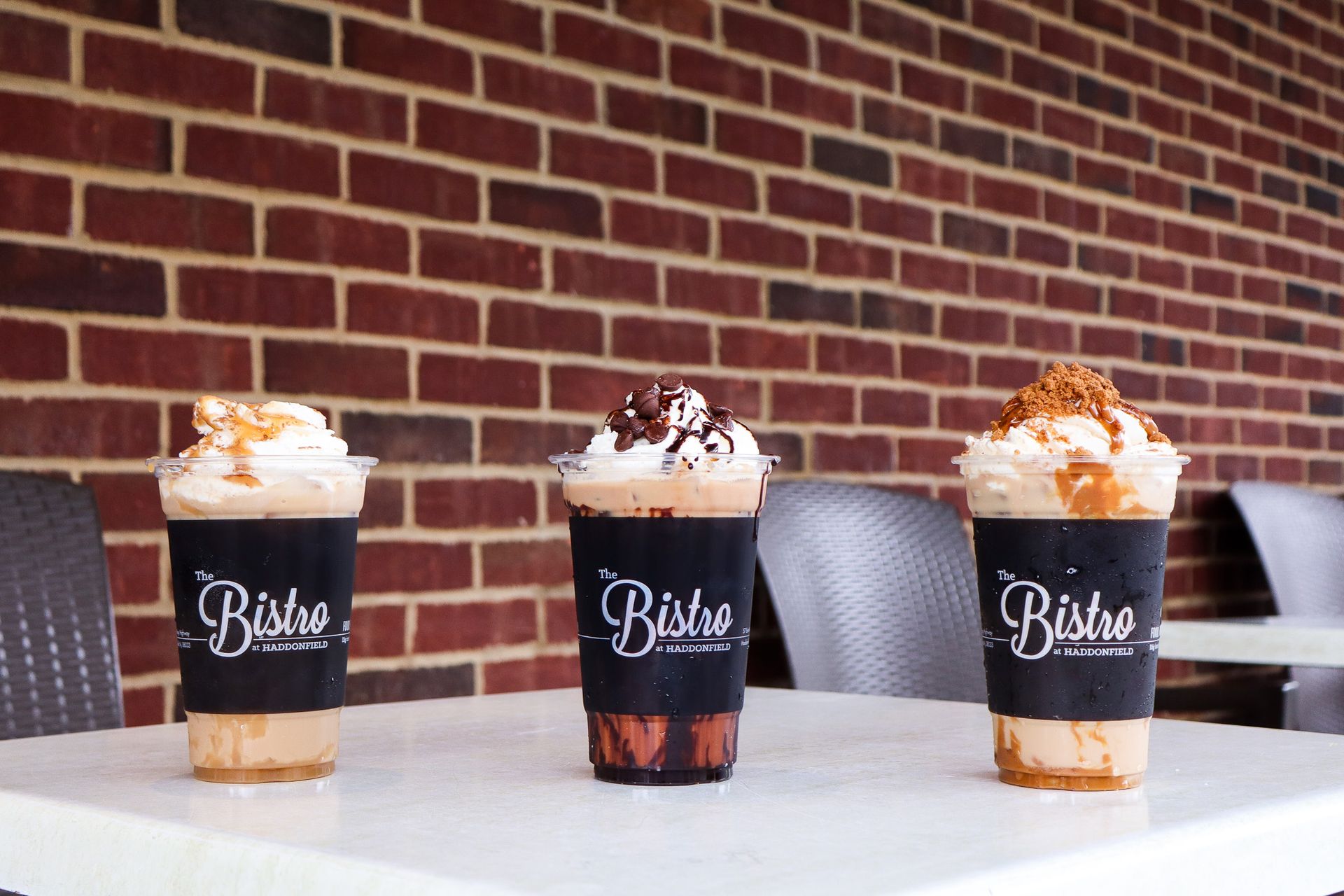 Three iced coffee drinks with whipped cream on a table against a brick wall.