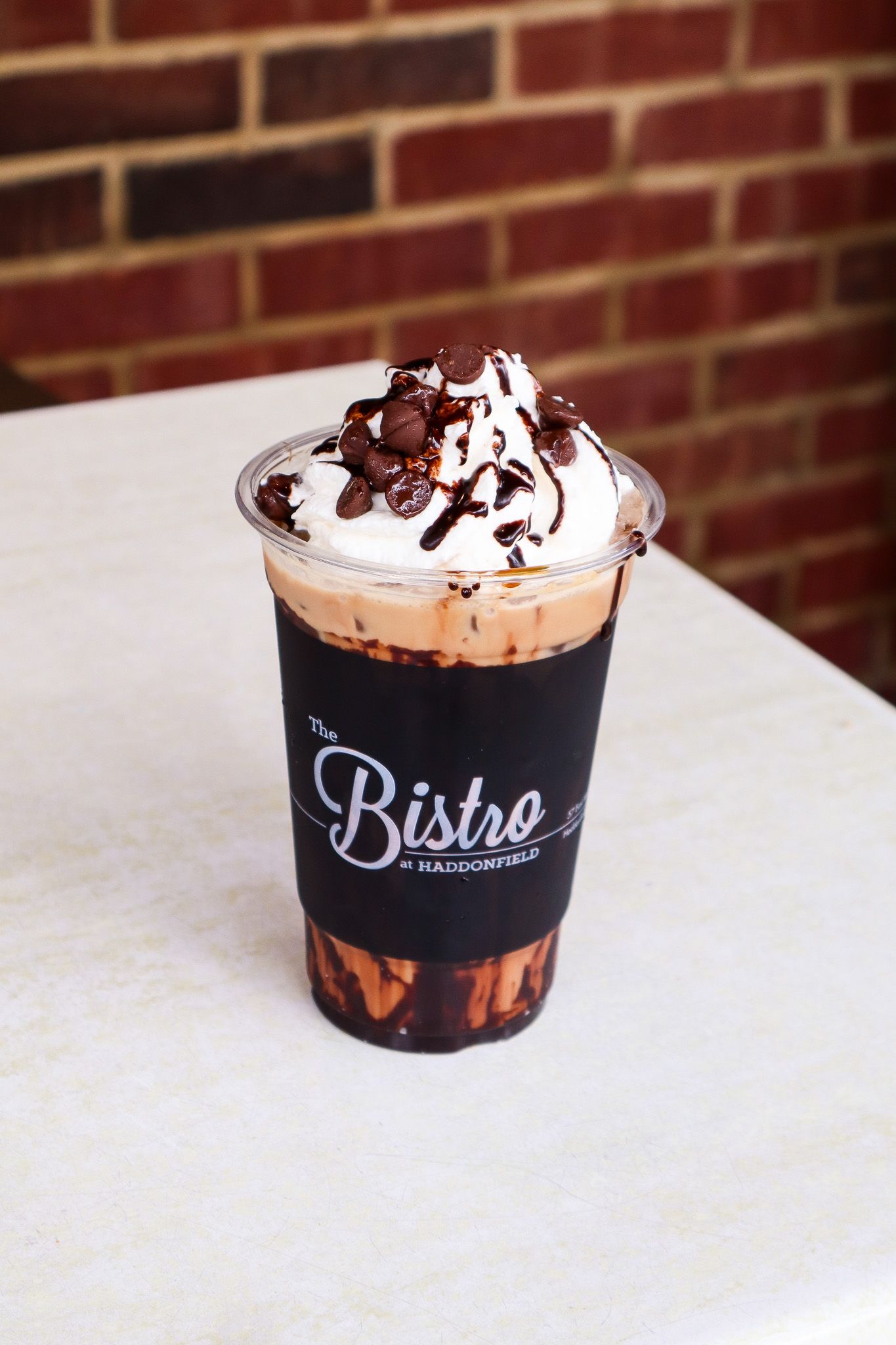 Chocolate milkshake topped with whipped cream and chocolate drizzle on a table, with a brick wall behind it