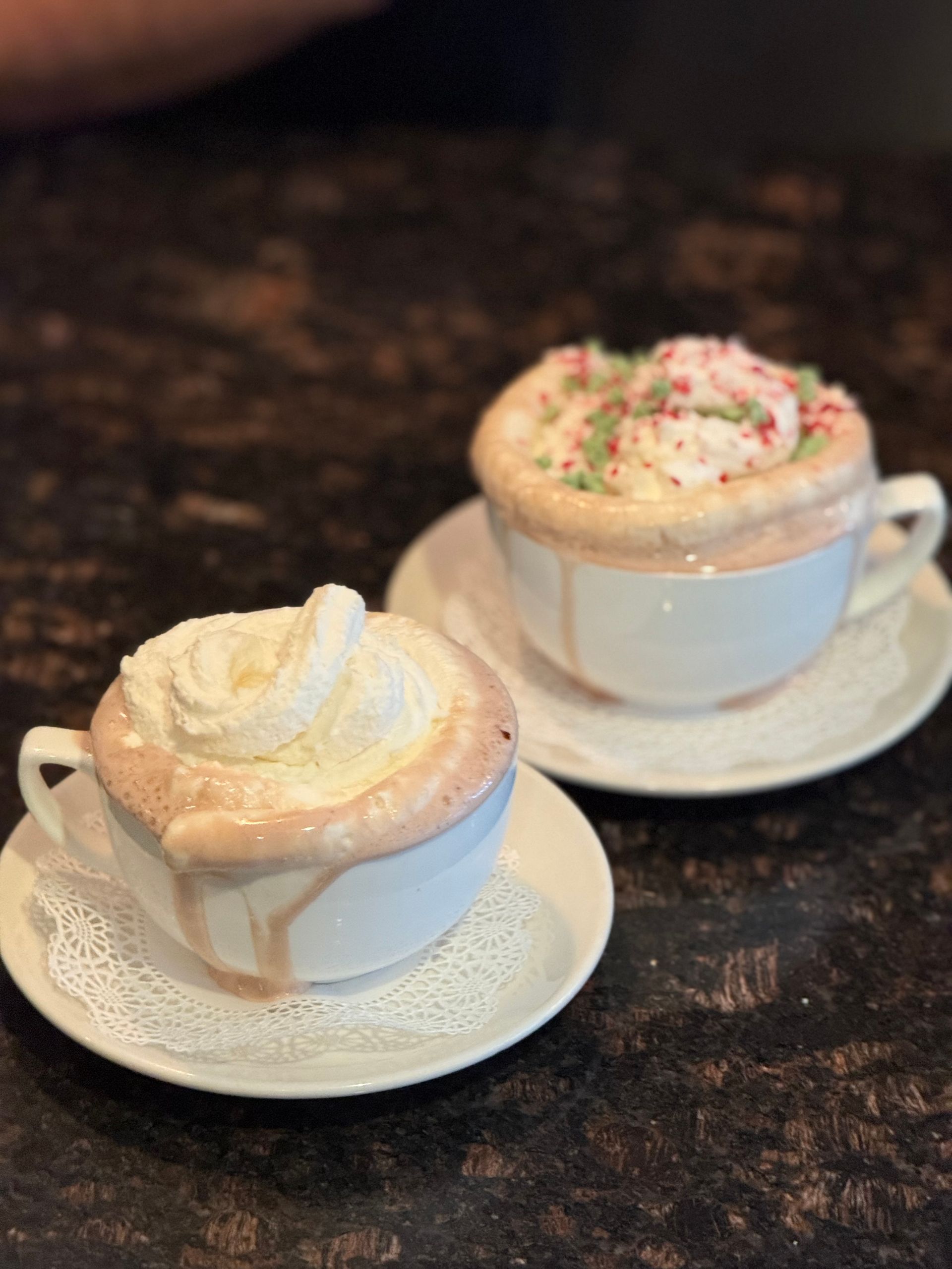 Two frothy coffee drinks on saucers, one plain and one topped with sprinkles, on a dark table