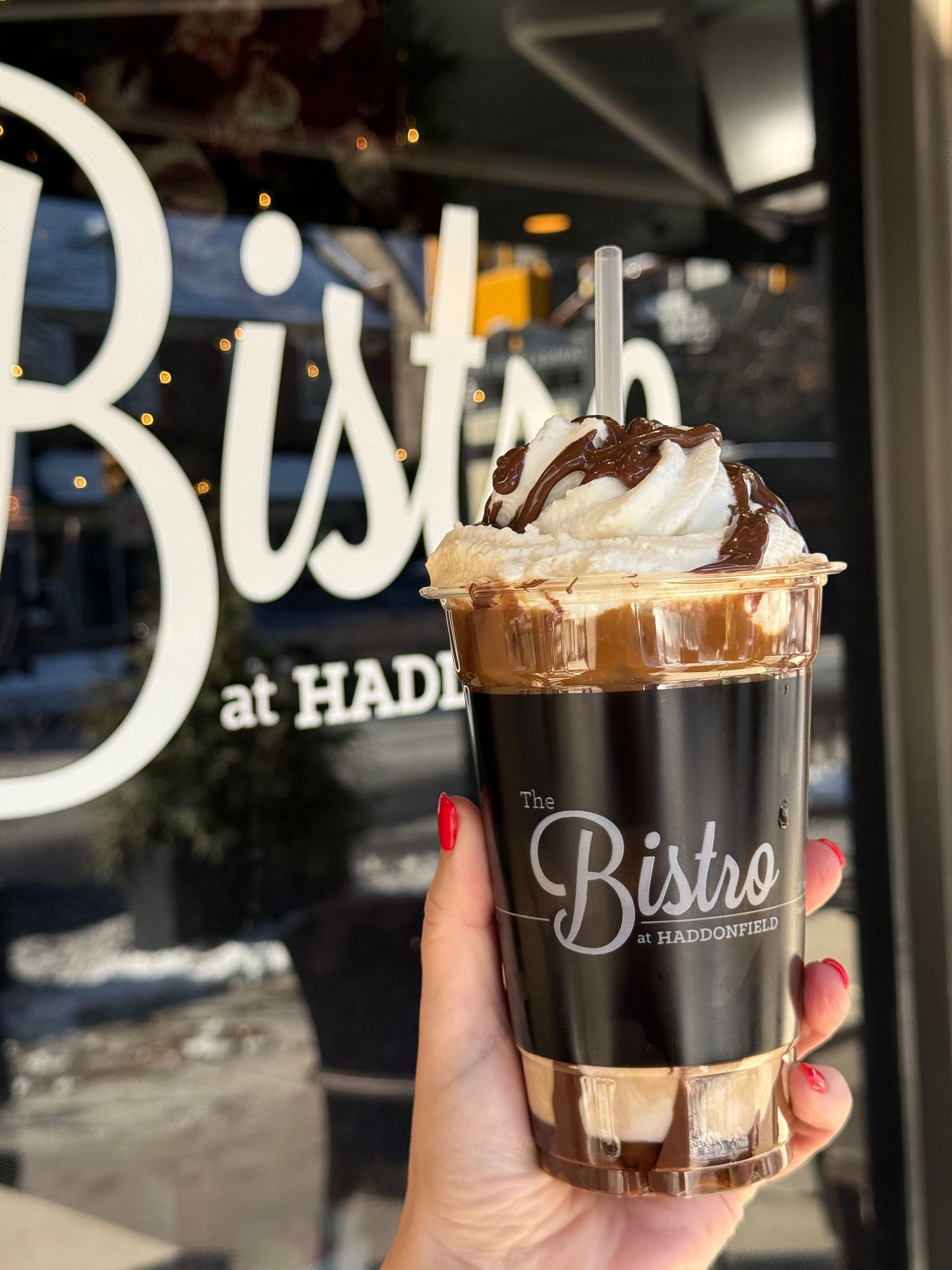 Hand holding a chocolate-drizzled whipped cream coffee cup outside Bistro cafe.