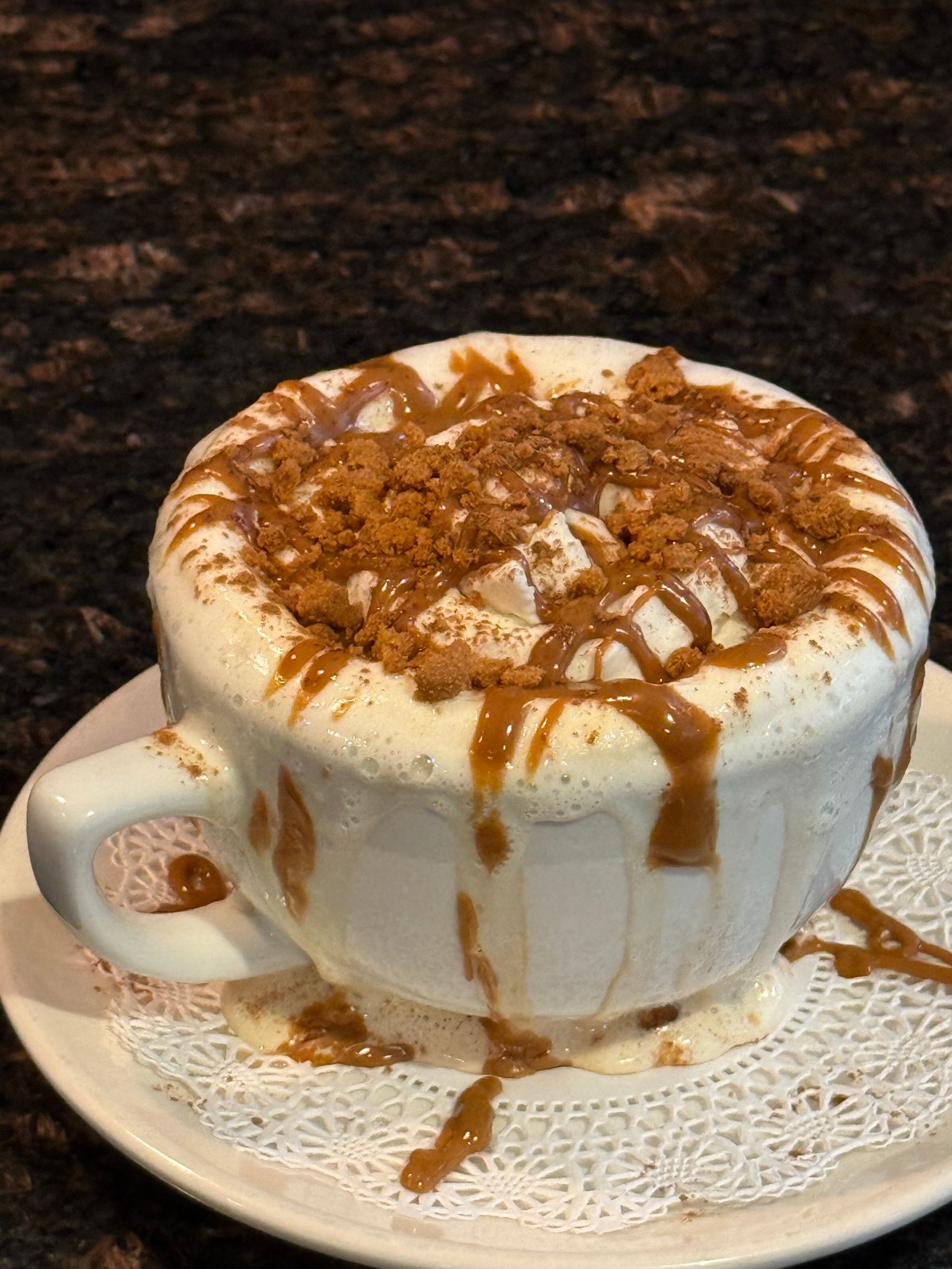 White mug of frothy coffee topped with caramel drizzle and cinnamon on a saucer with lace doily