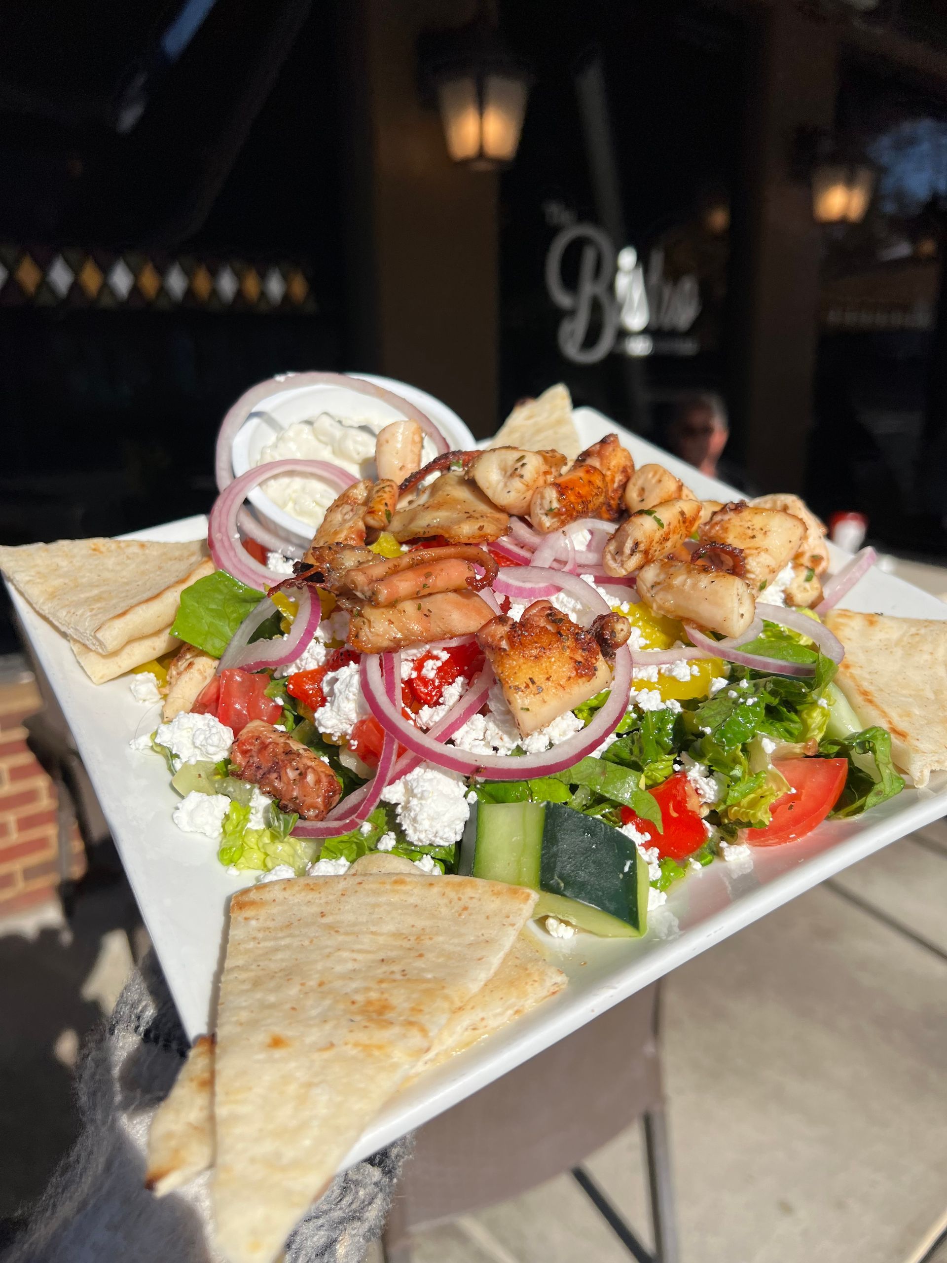 Greek salad with grilled chicken and pita on a square plate outdoors at a restaurant