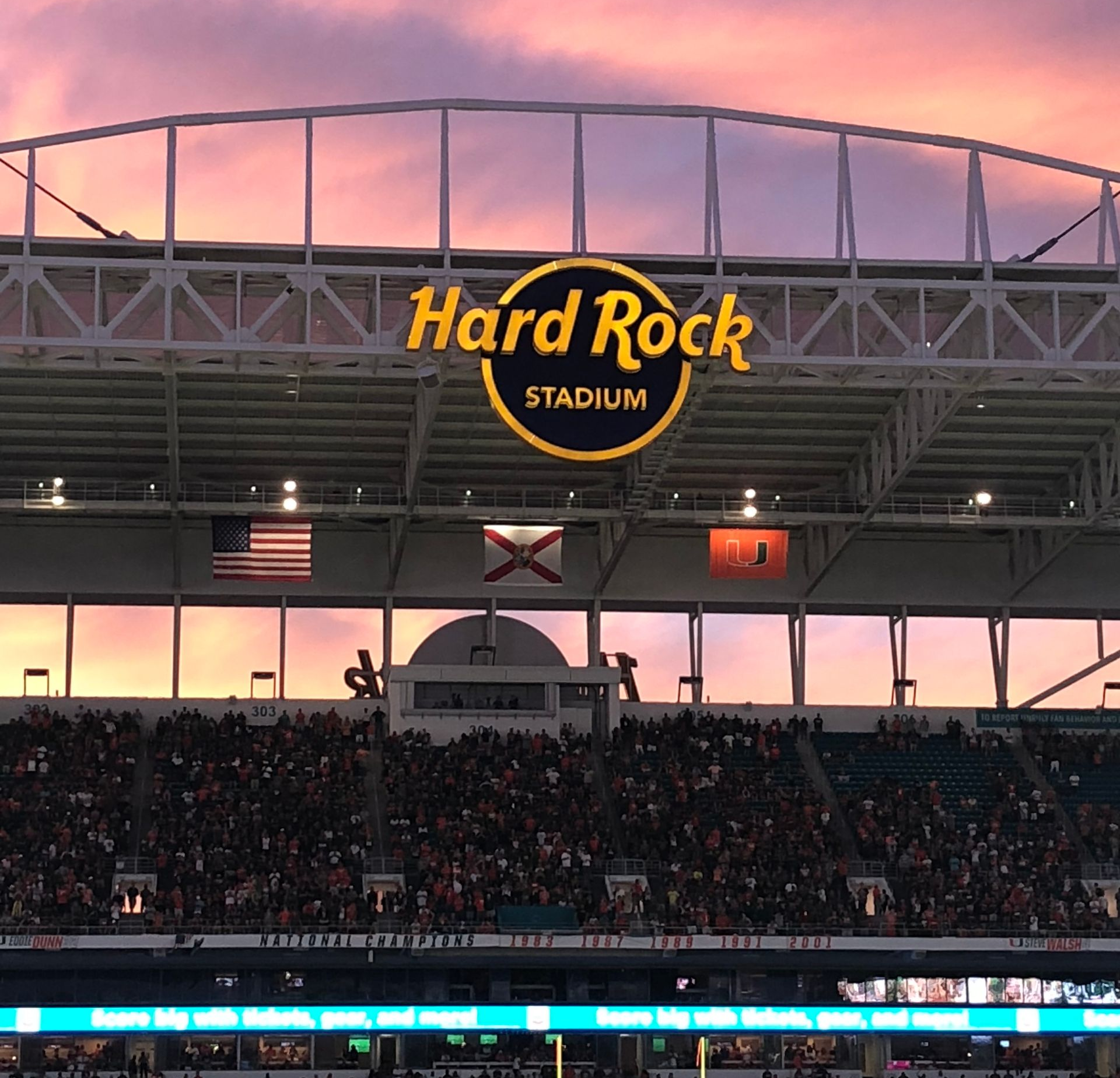 Hard Rock Stadium sign at sunset over a packed stadium with American and Florida flags.