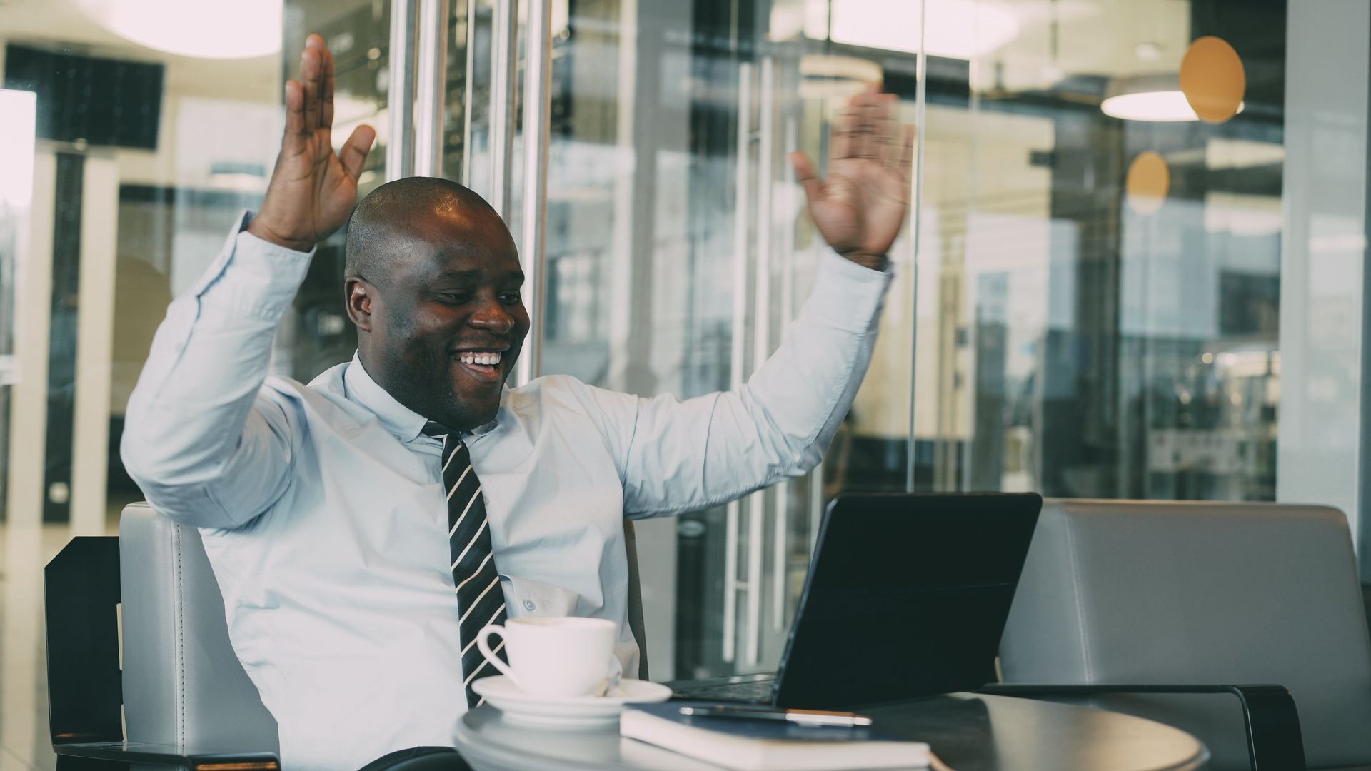 Man at office desk cheering with both hands raised beside a laptop and coffee cup