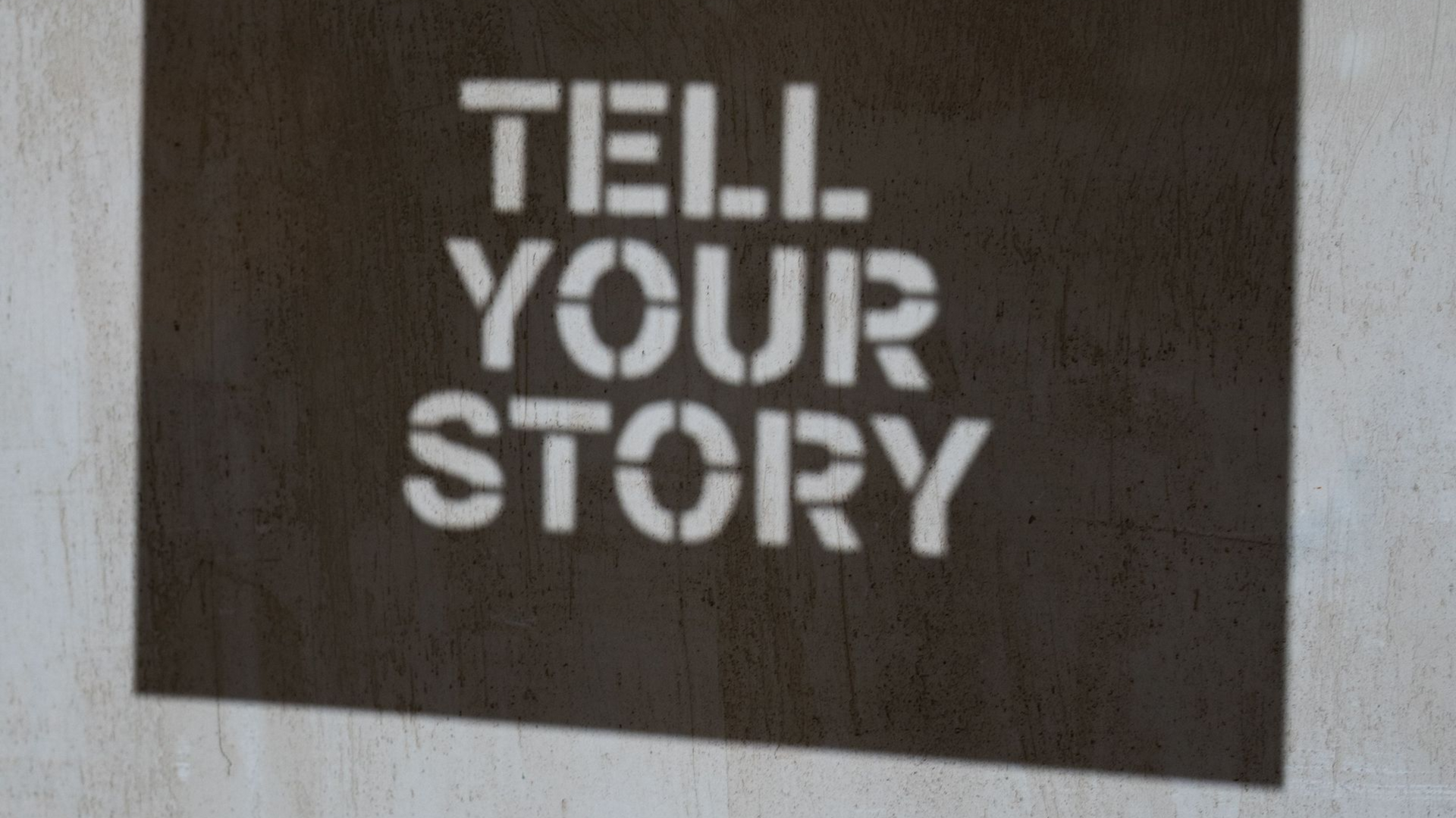 Dark rectangular sign with white stencil text reading “TELL YOUR STORY” on a light wall.