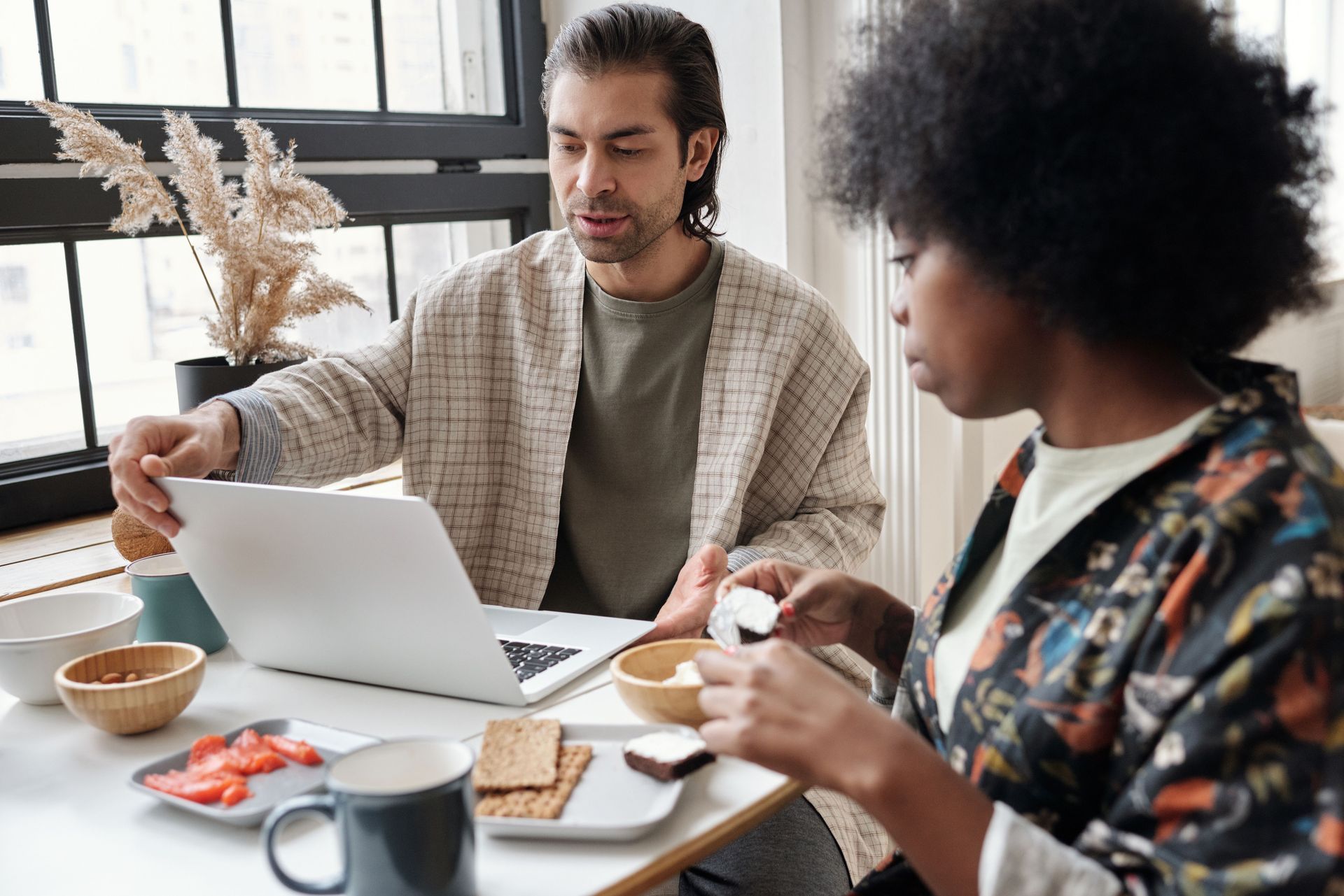 Two people working at a table with a laptop, bowls, bread, and coffee in a bright office or café