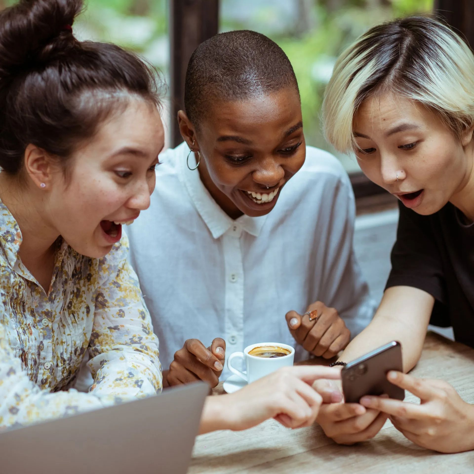 Three diverse women excitedly look at a phone, one points, with surprised expressions.