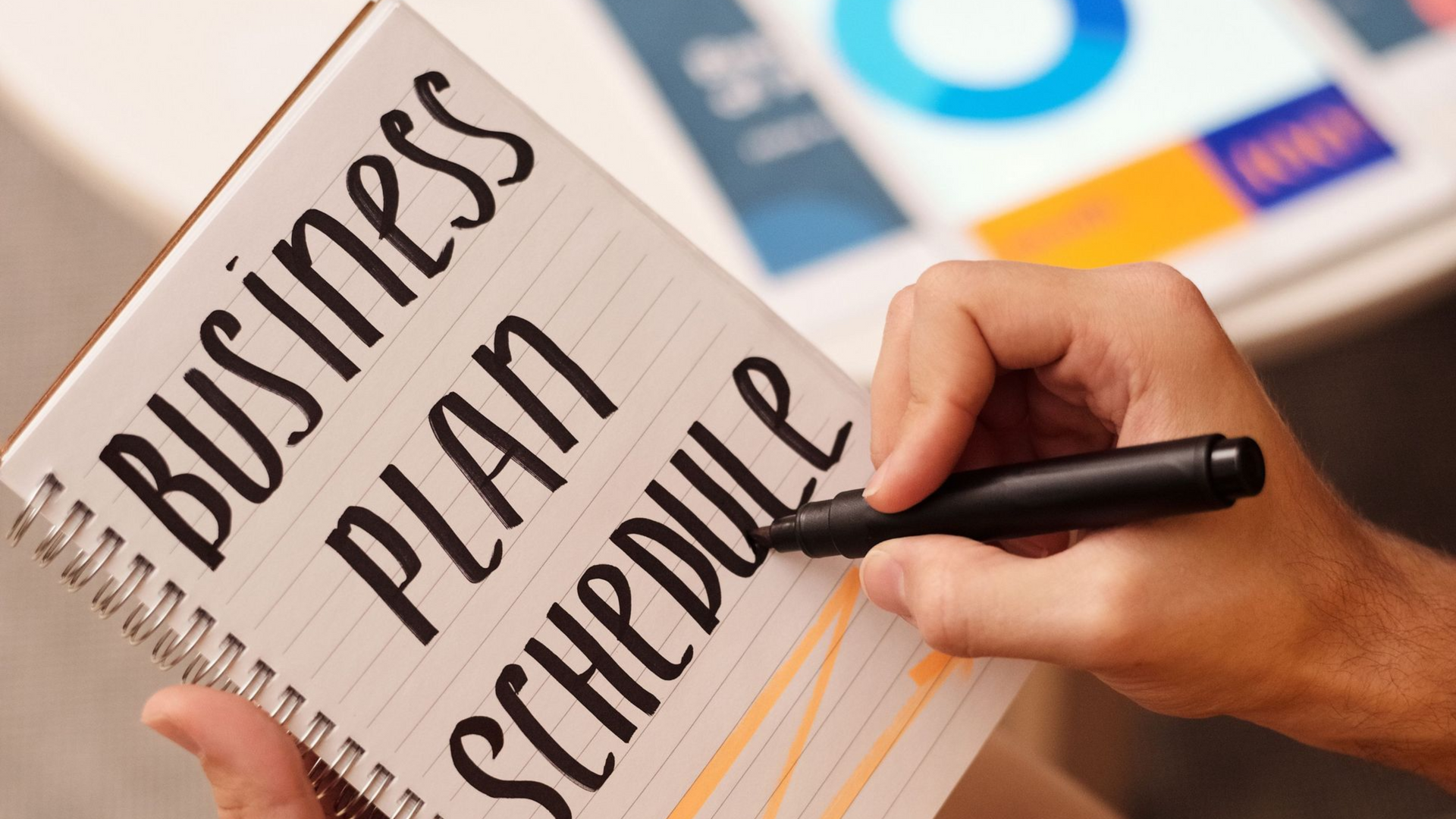 Handwriting a business plan schedule in a notebook beside a laptop with charts