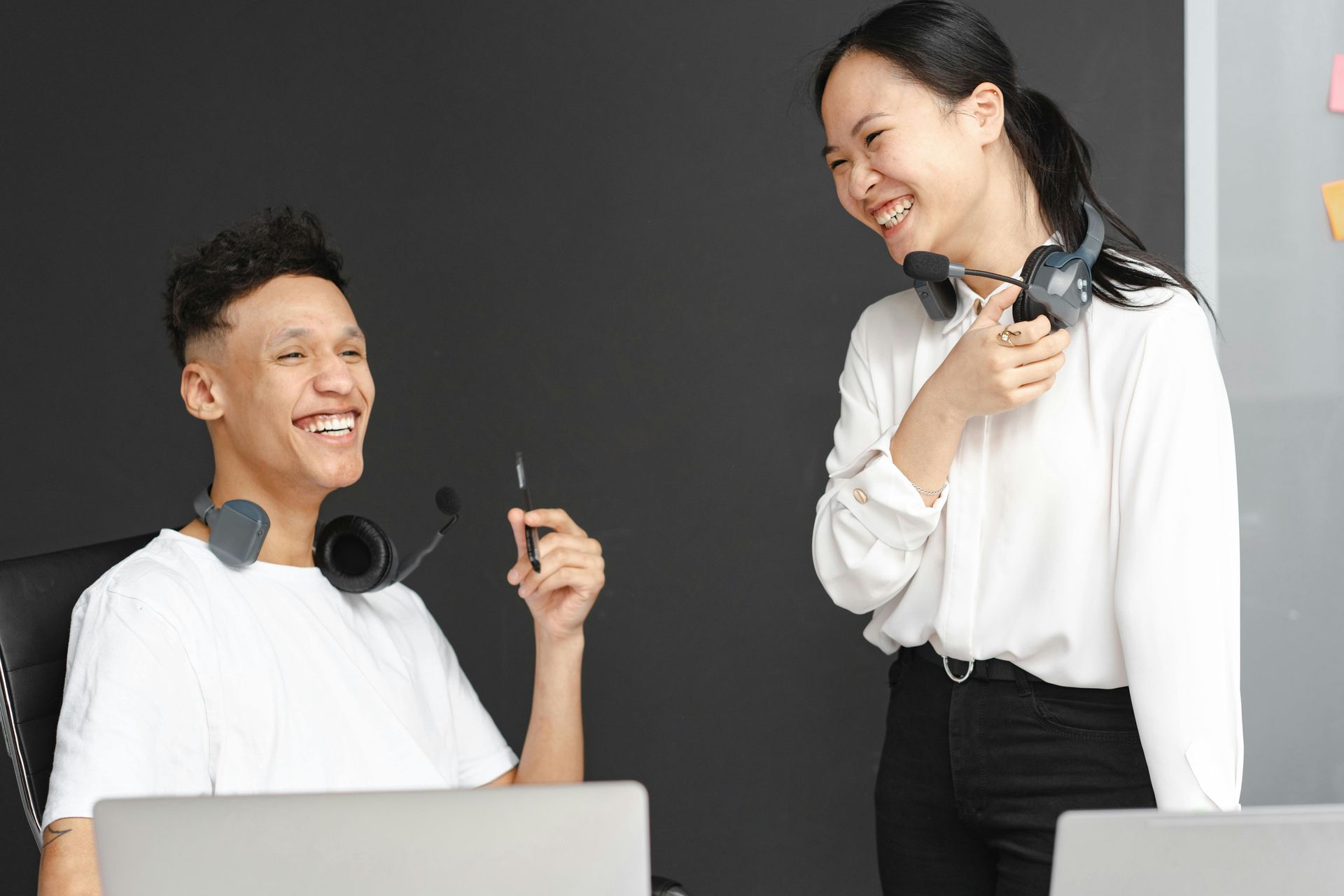 Two coworkers smiling at a desk, talking with headphones around their necks in a bright office