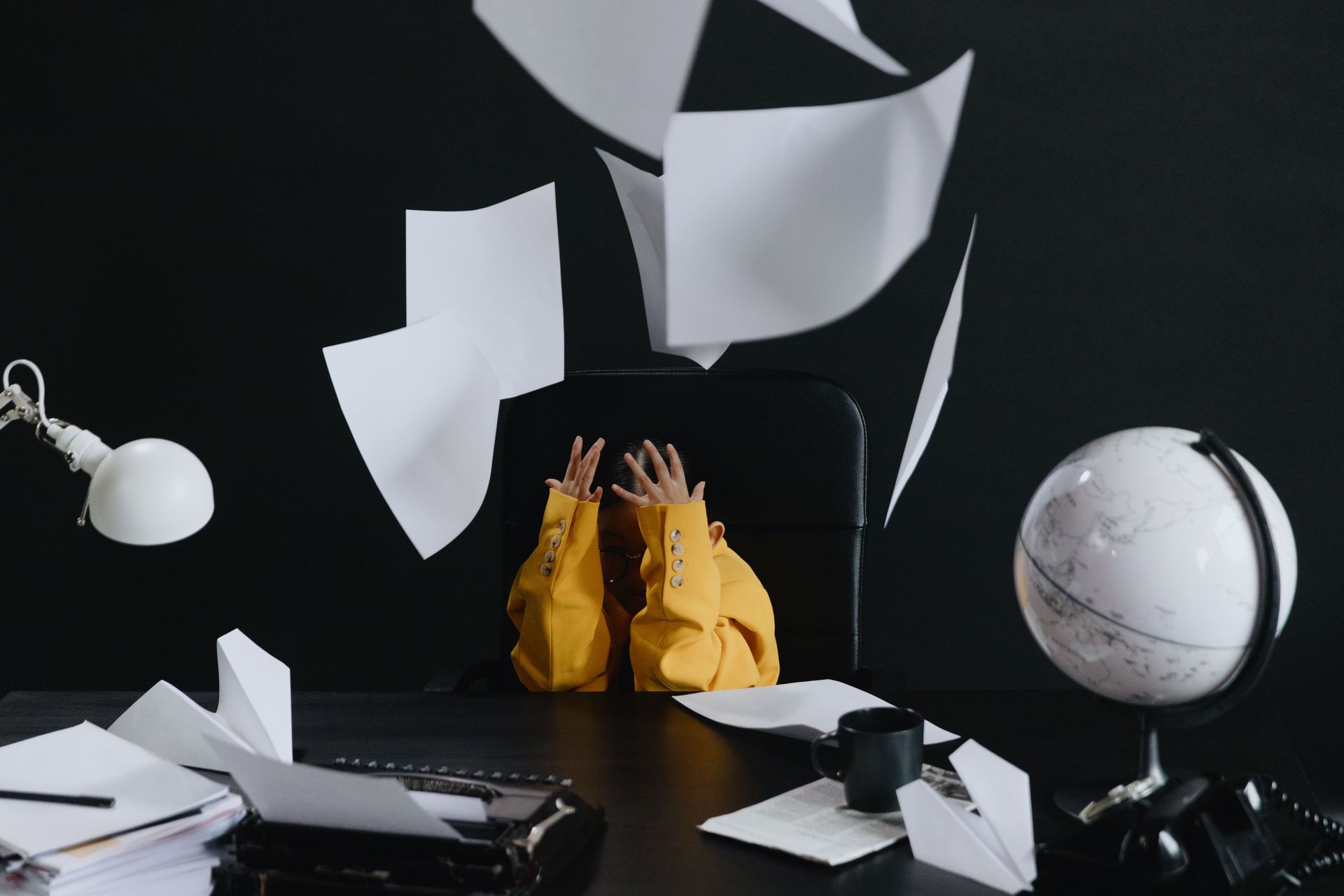 Person in yellow among flying papers, with a globe and desk lamp on a dark desk