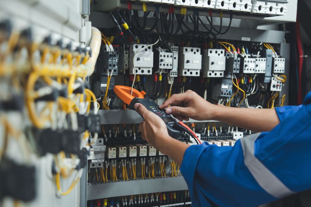 Electrician in blue uniform using a multimeter to inspect electrical wiring in a control panel.