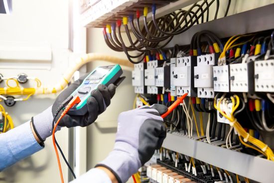 Electrician in blue uniform using a multimeter to inspect electrical wiring in a control panel.