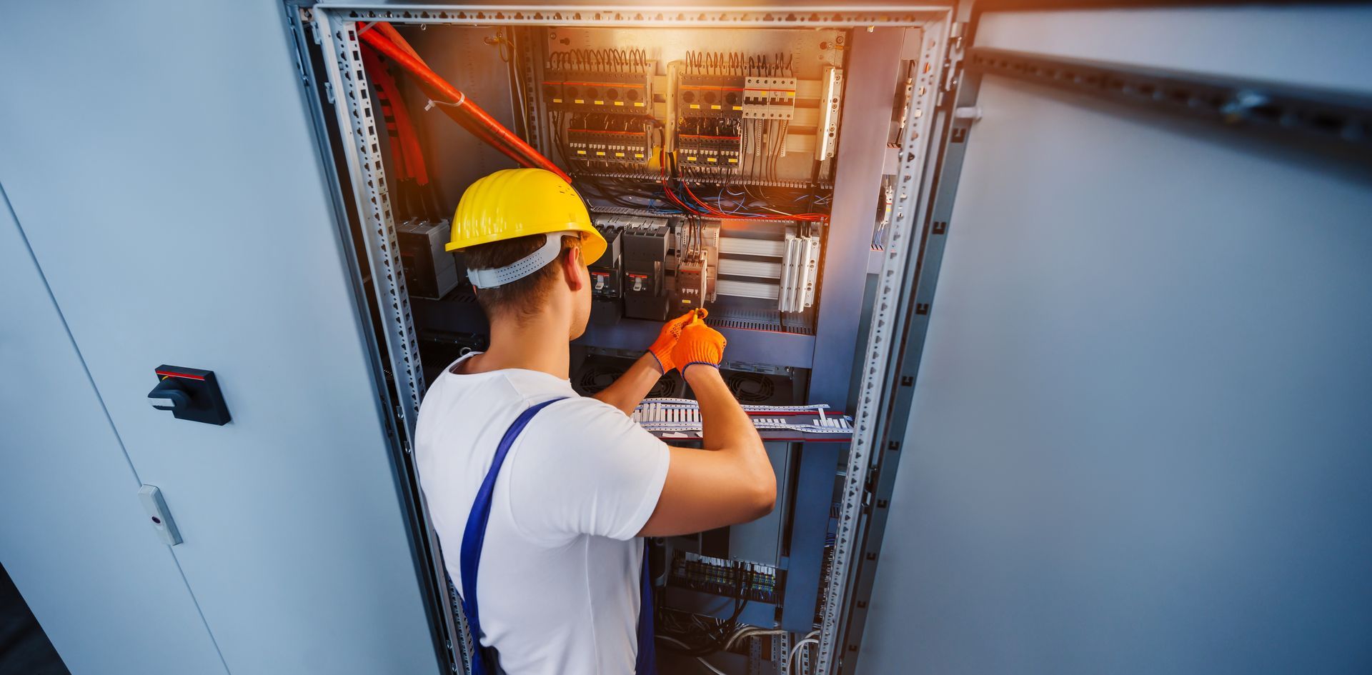 Electrician working on electrical panel, wearing yellow hard hat and gloves.