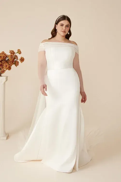 Bride in white off-the-shoulder gown, smiling in a studio setting with flowers and a small pillar.