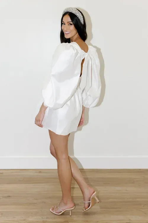 Woman in white dress with puffy sleeves and open back, wearing a pearl headband and heels, smiling.
