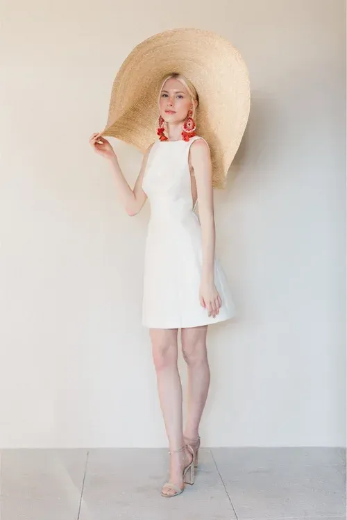 Woman in white dress, oversized straw hat, red earrings, and nude heels, standing in front of a cream wall.