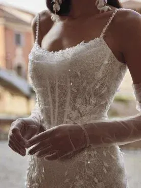 Woman in a white sequined wedding dress with spaghetti straps and long sheer gloves, outdoors.