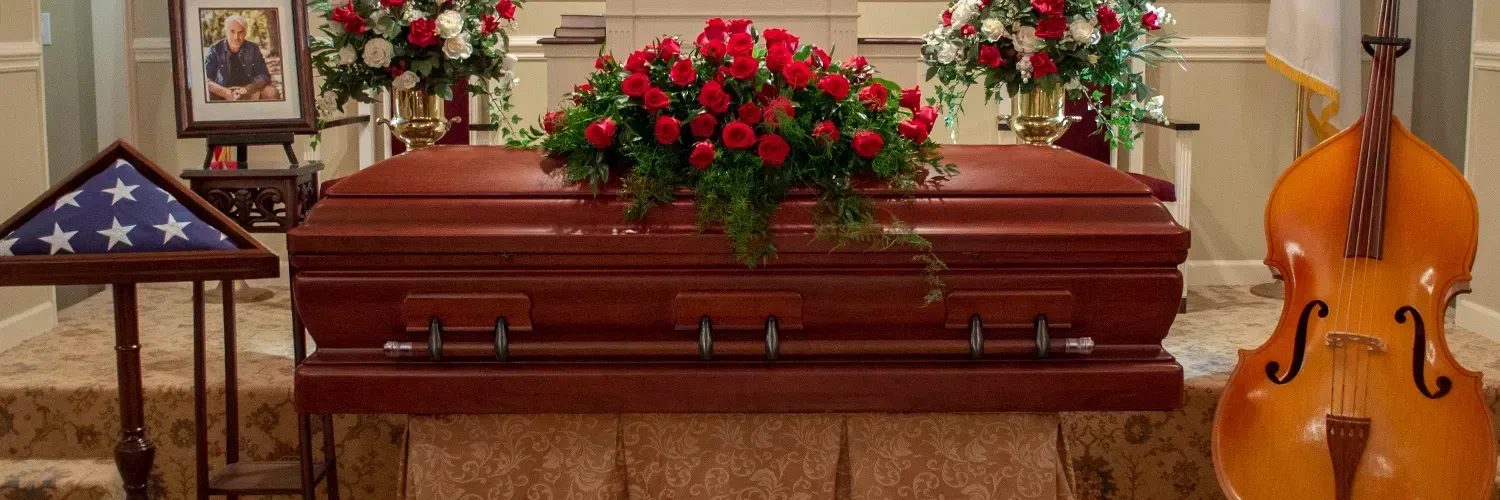 Wooden casket with red roses, flanked by an American flag and an upright bass in a funeral setting