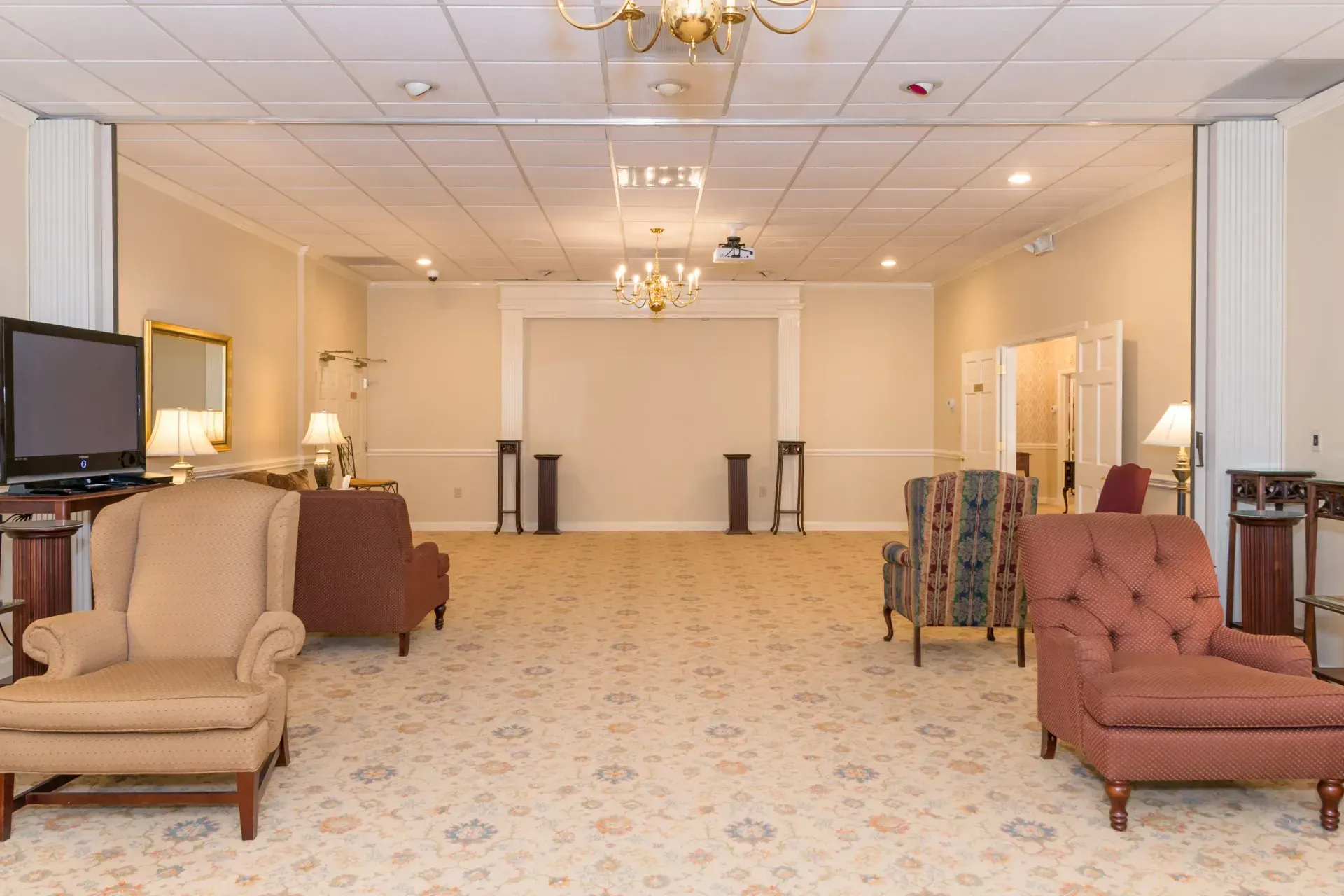 Elegant banquet hall with beige carpet, chandeliers, and upholstered chairs arranged around a spacious open floor
