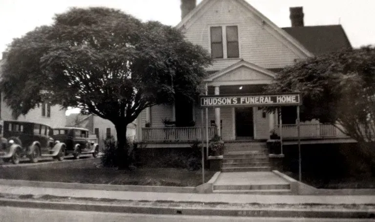 Historic funeral home with porch and cars parked outside on a tree-lined street