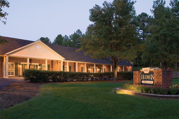 Single-story building at dusk with lit windows and a stone sign by a green lawn.