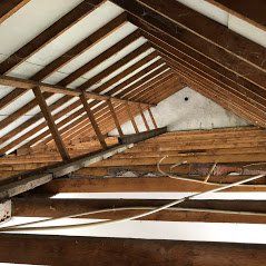 Een onafgewerkte zolder met zichtbare houten balken, witte dakpanelen en zichtbare elektrische bedrading.