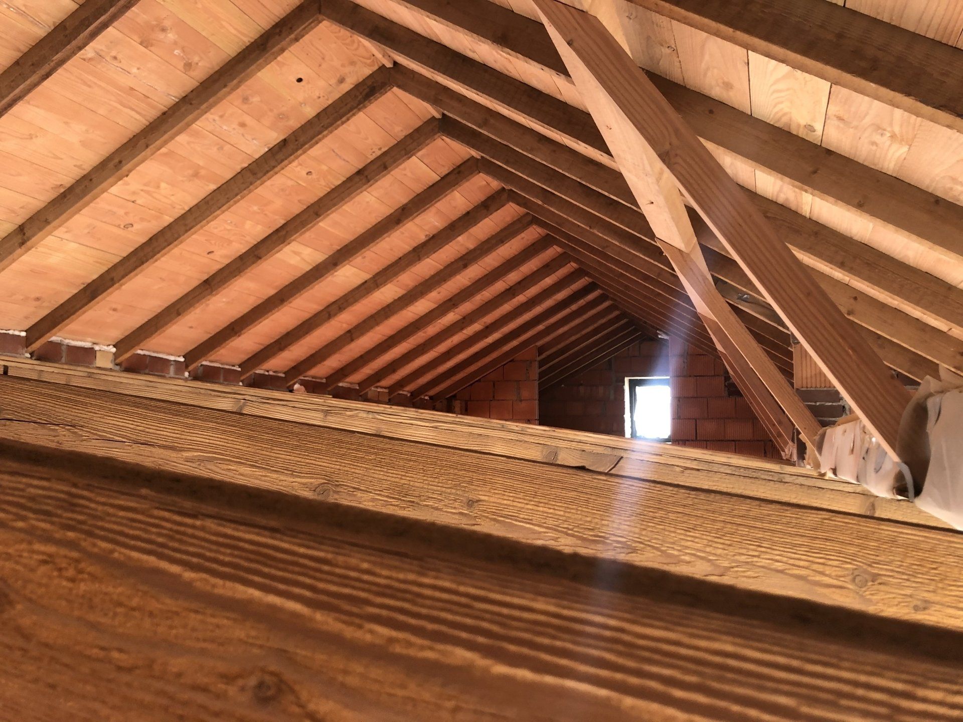 Een zolderinterieur in aanbouw, met houten spanten, balken en een klein raam in de bakstenen muur in de verte.