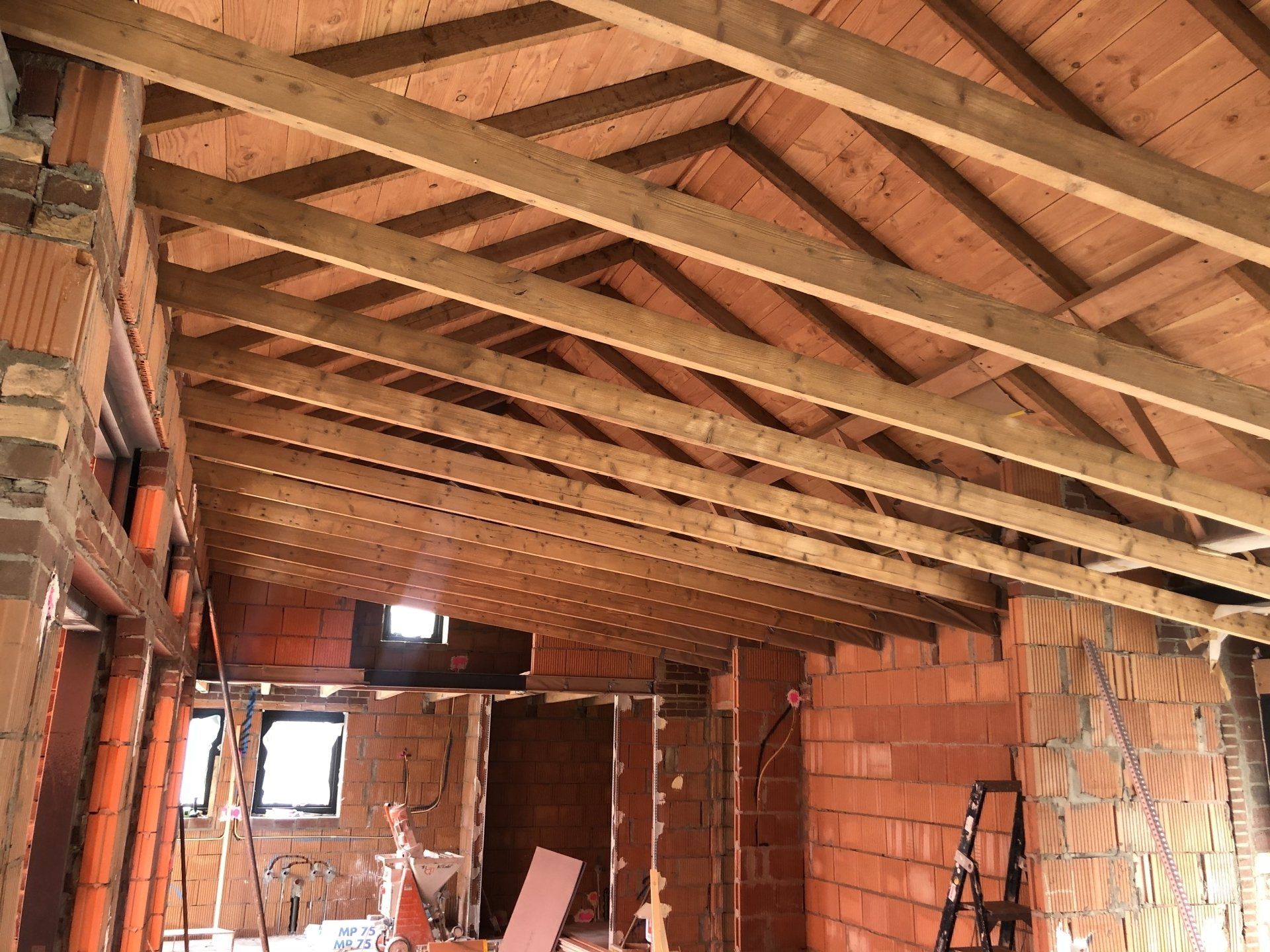 Interieur van een bouwplaats met een houten dakconstructie, zichtbare bakstenen muren en open raamkozijnen.