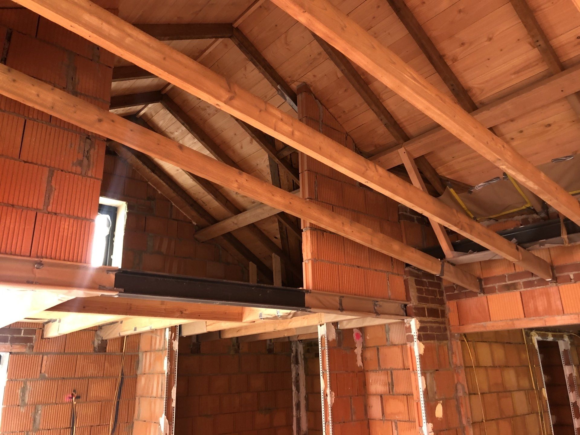 Interieurfoto van een bouwplaats met een onafgewerkte bakstenen muur, zichtbare houten dakbalken en stalen steunconstructie.