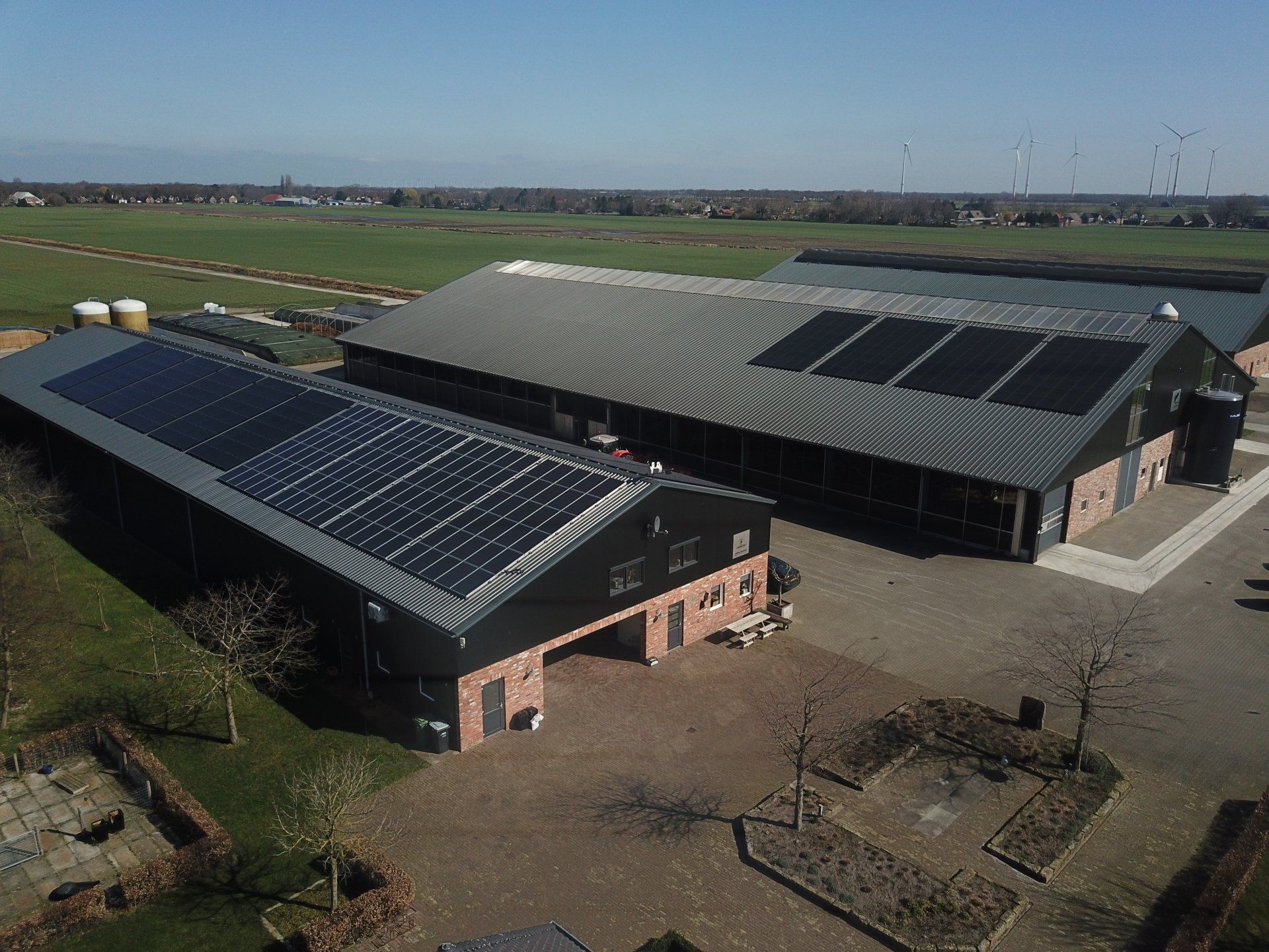 Een luchtfoto van twee boerderijgebouwen met zonnepanelen op hun metalen daken, gelegen in een vlak, landelijk landschap.