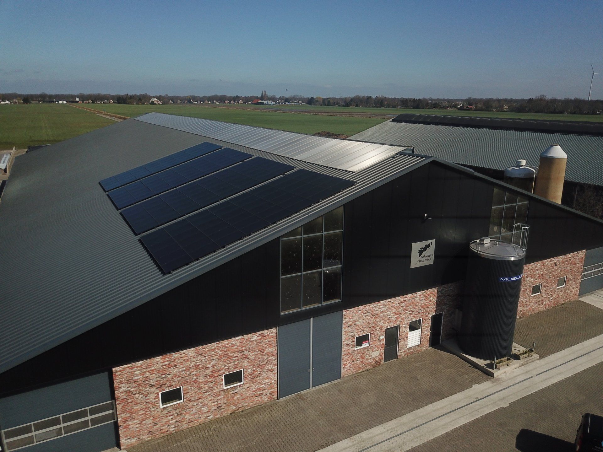 Een modern agrarisch gebouw met een zwart metalen dak, zonnepanelen en rode bakstenen muren, gelegen in een landelijk landschap.