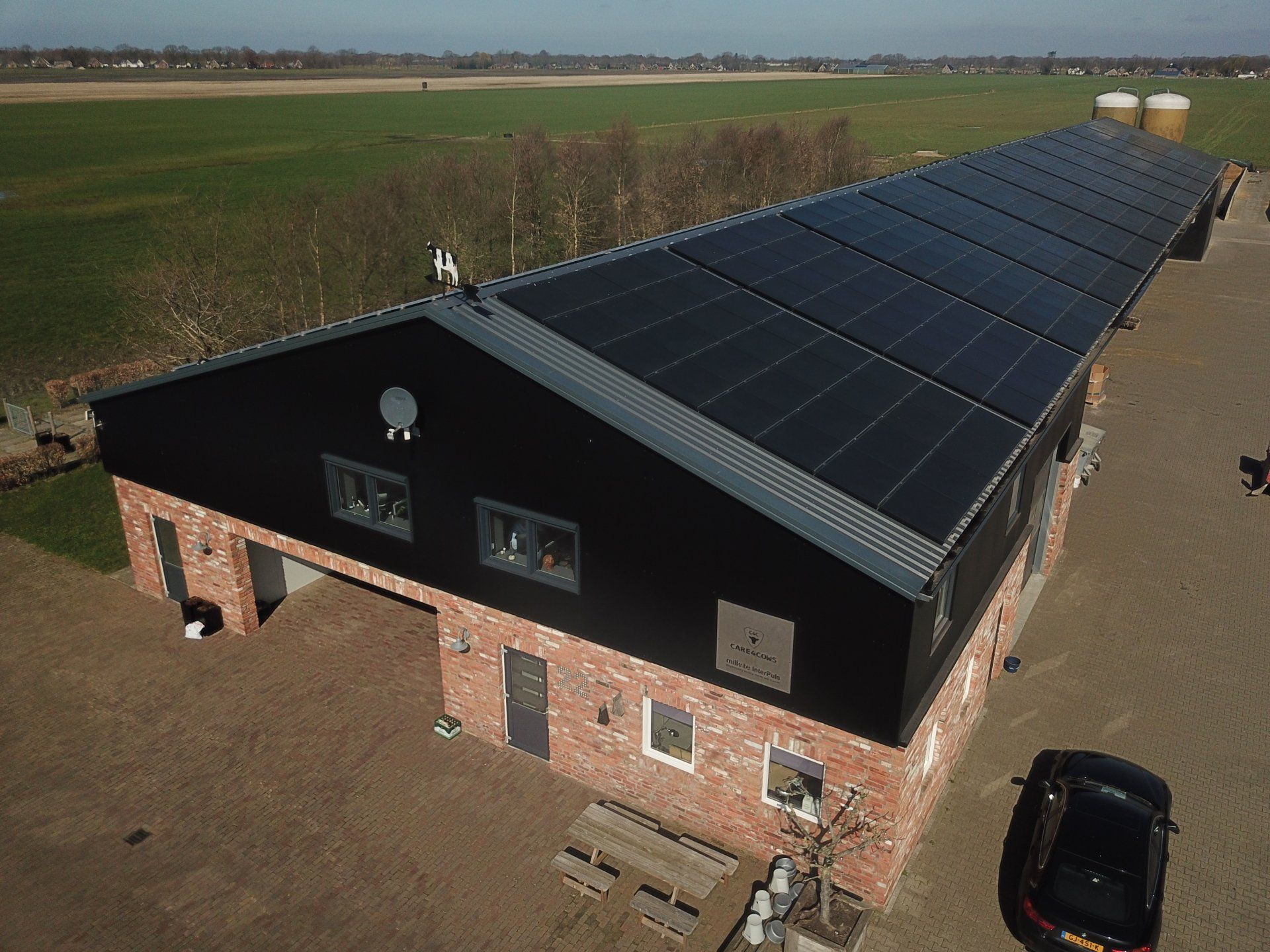 Een lang bakstenen gebouw met een zwart metalen dak en een grote installatie van zonnepanelen op het dak, gelegen in een landelijk landschap.
