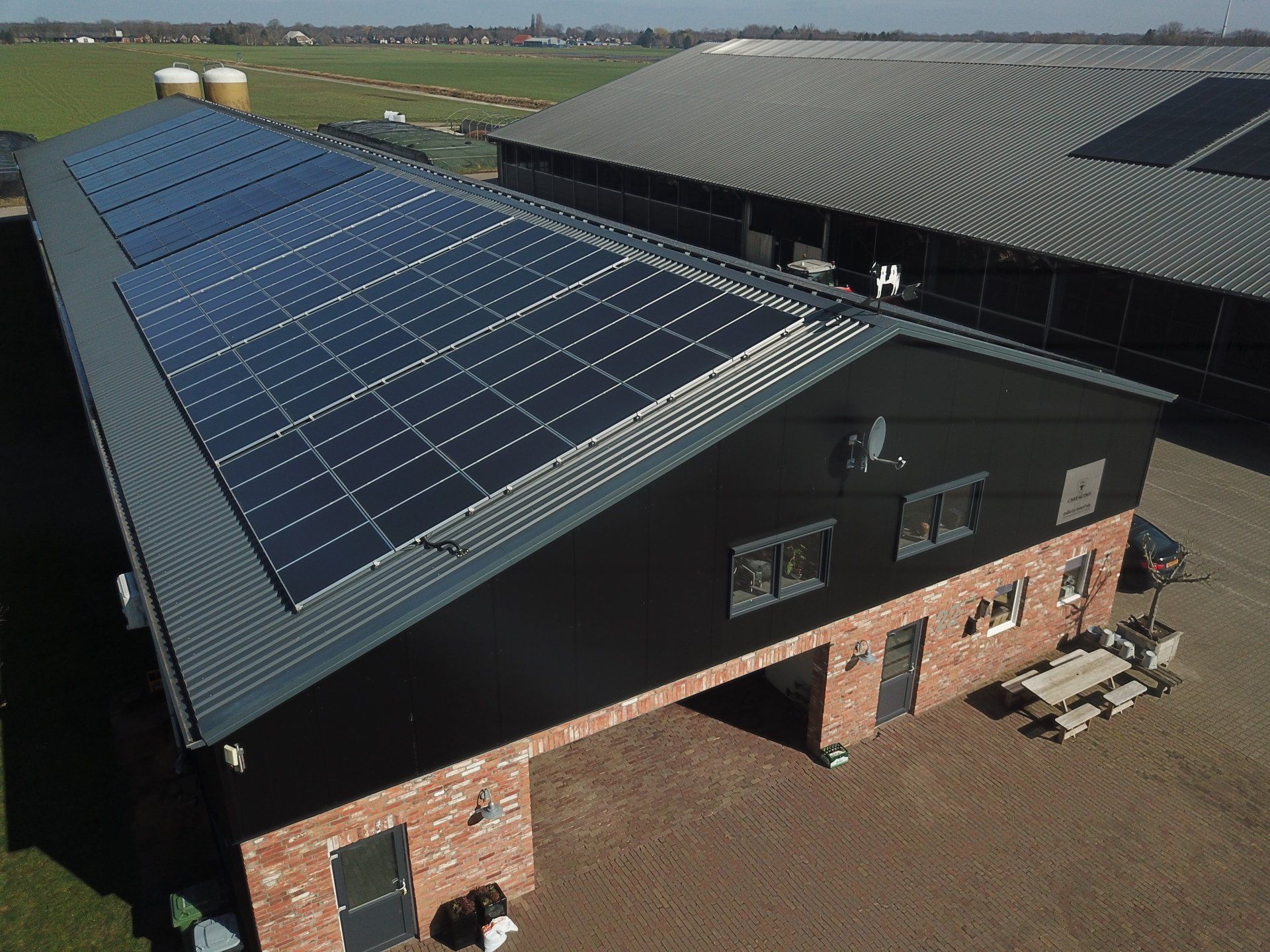Luchtfoto van een boerderijgebouw met een groot zonnepanelenveld op het dak, naast een grotere schuur, onder een heldere hemel.