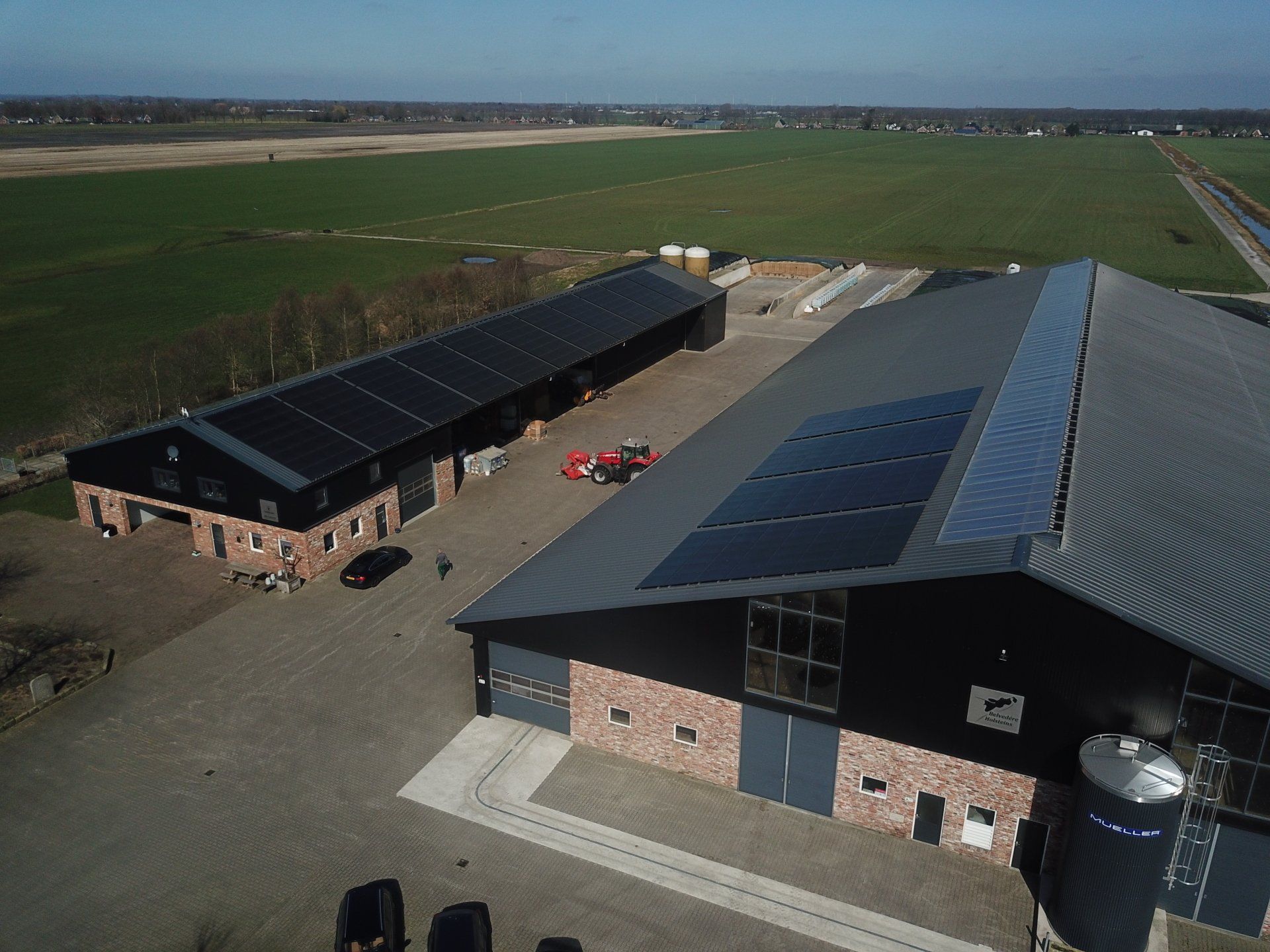 Luchtfoto van een boerderij met twee grote agrarische gebouwen met zonnepanelen, een verhard erf en uitgestrekte groene velden.