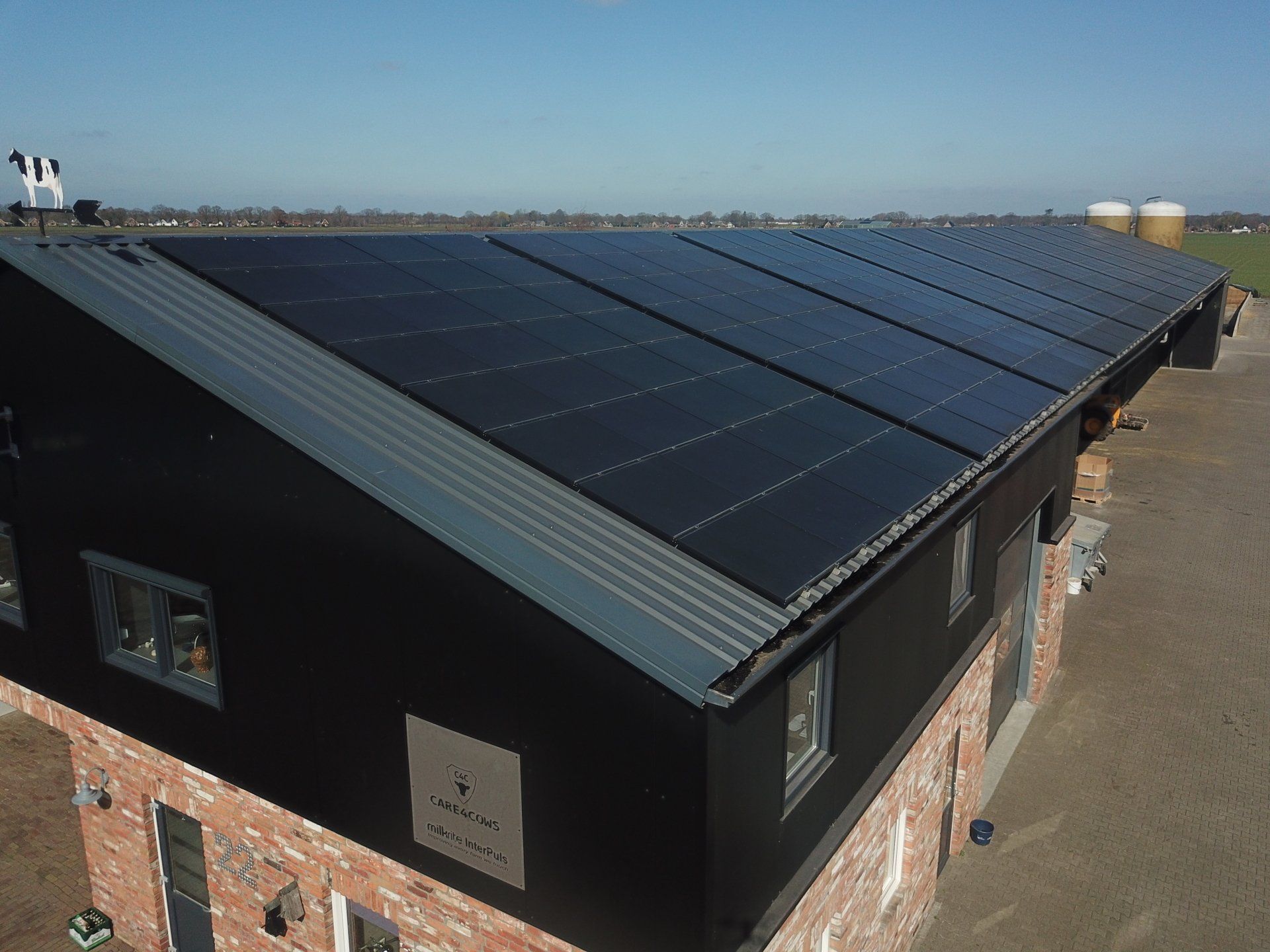 Een luchtfoto van een boerderijgebouw met een donker dak dat volledig bedekt is met zonnepanelen, gelegen in een landelijk, zonnig landschap.