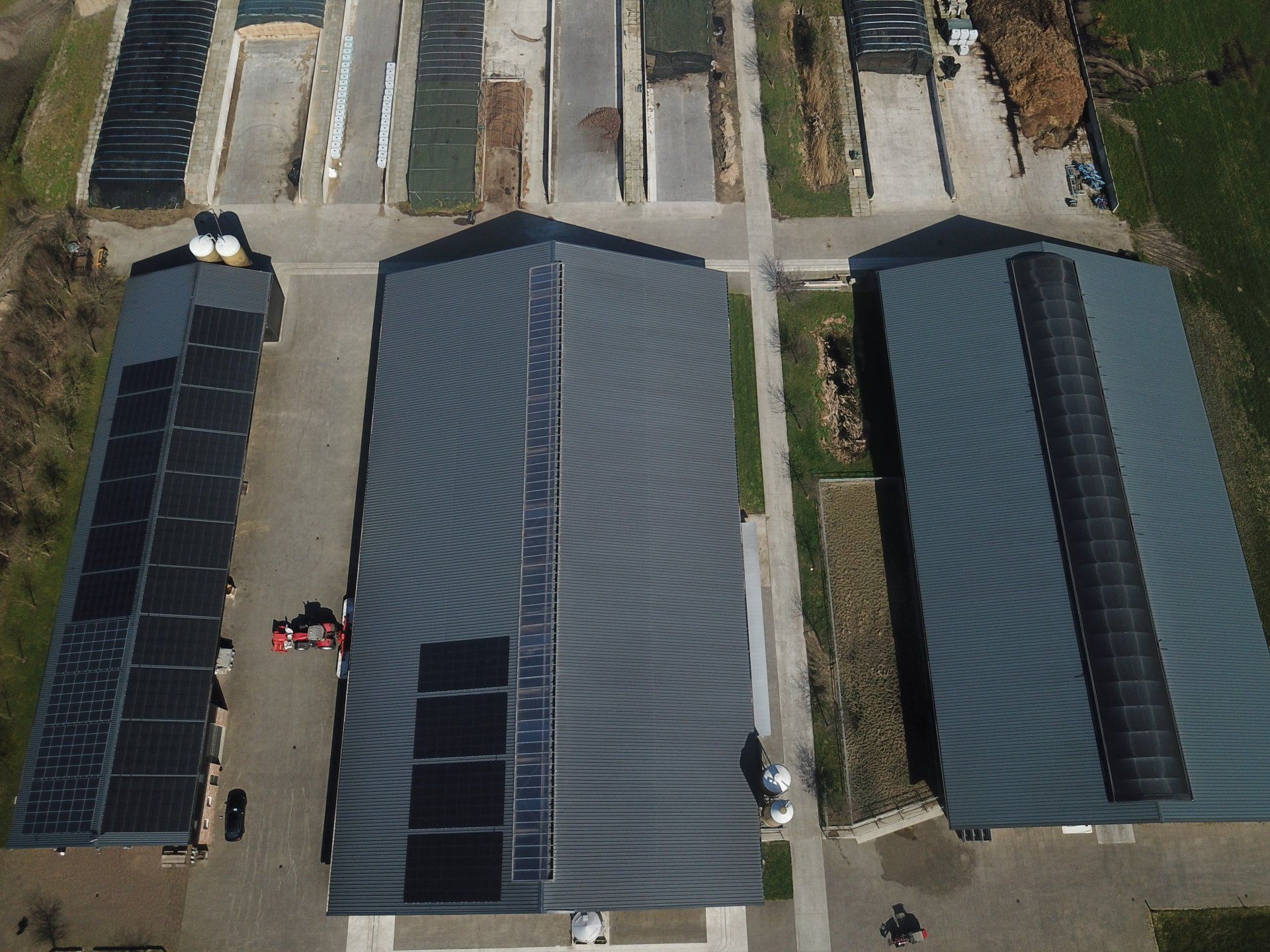 Luchtfoto van drie gebouwen met grijze daken en aangrenzende rechthoekige buitenopslagruimtes op een verhard industrieterrein.