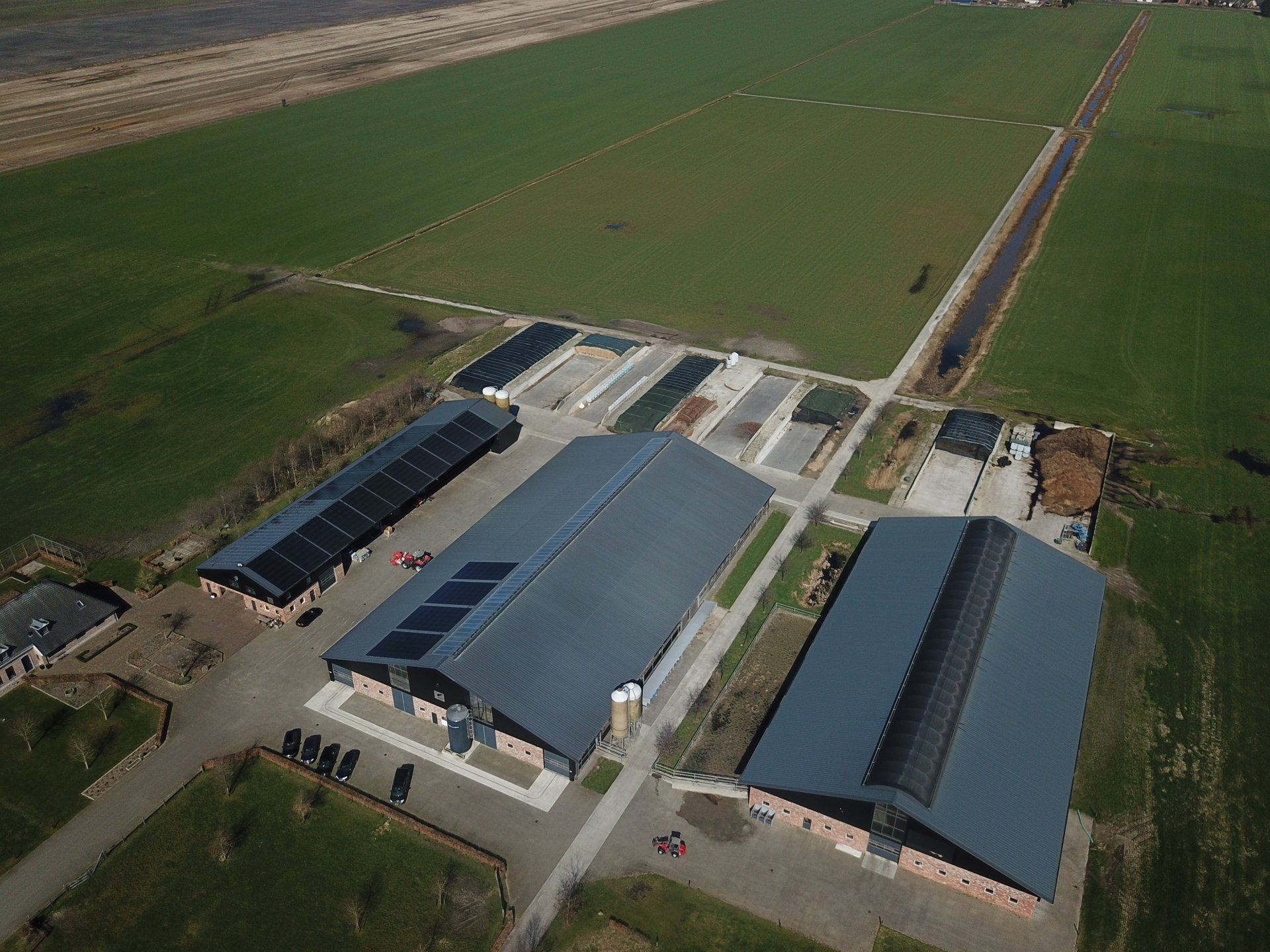 Een luchtfoto van een agrarisch complex met drie grote industriële gebouwen, omgeven door groene velden.