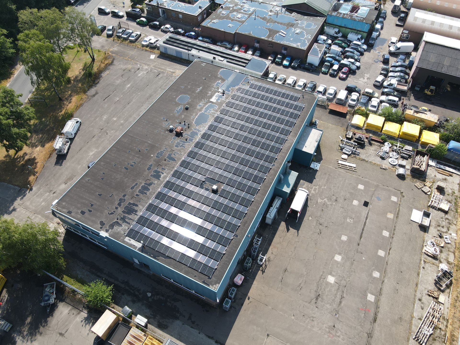 Luchtfoto van een rechthoekig industrieel gebouw met een grote zonnepaneleninstallatie op het dak, omgeven door parkeerplaatsen.