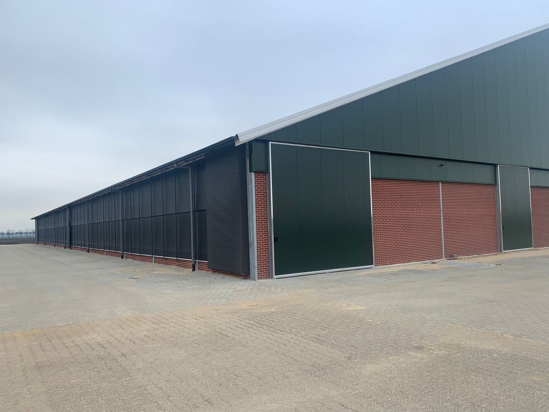 Een groot, langgerekt agrarisch gebouw met groene metalen gevelbekleding en bakstenen ventilatiepanelen op een grindterrein onder een grijze hemel.