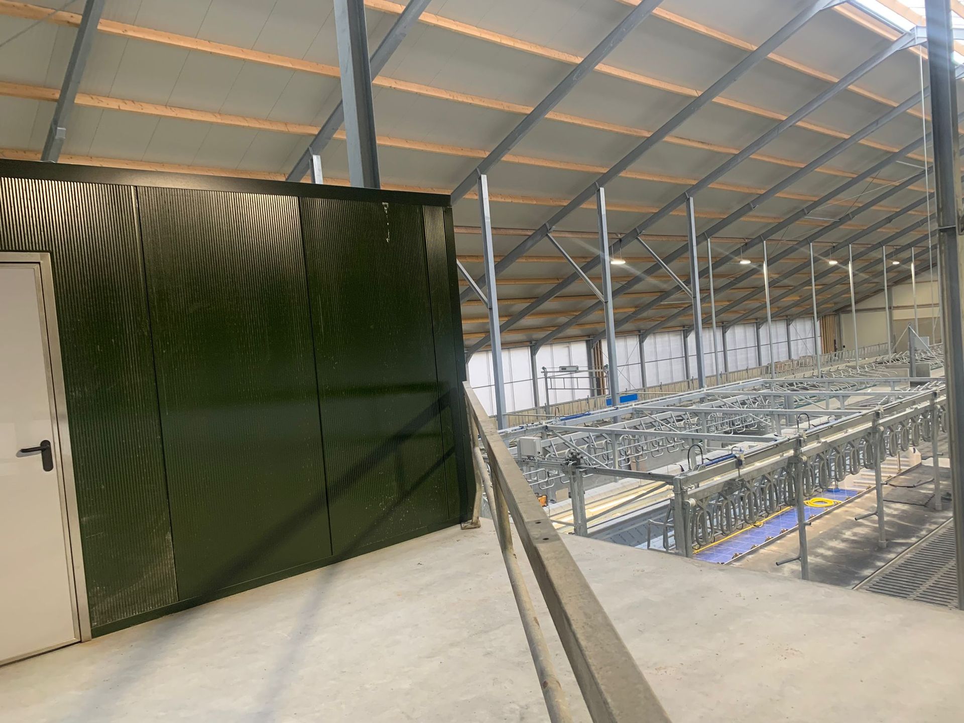 Een verhoogde loopbrug biedt uitzicht op een ruime melkveestal met metalen omheining en stallen onder een hoog, schuin dak.