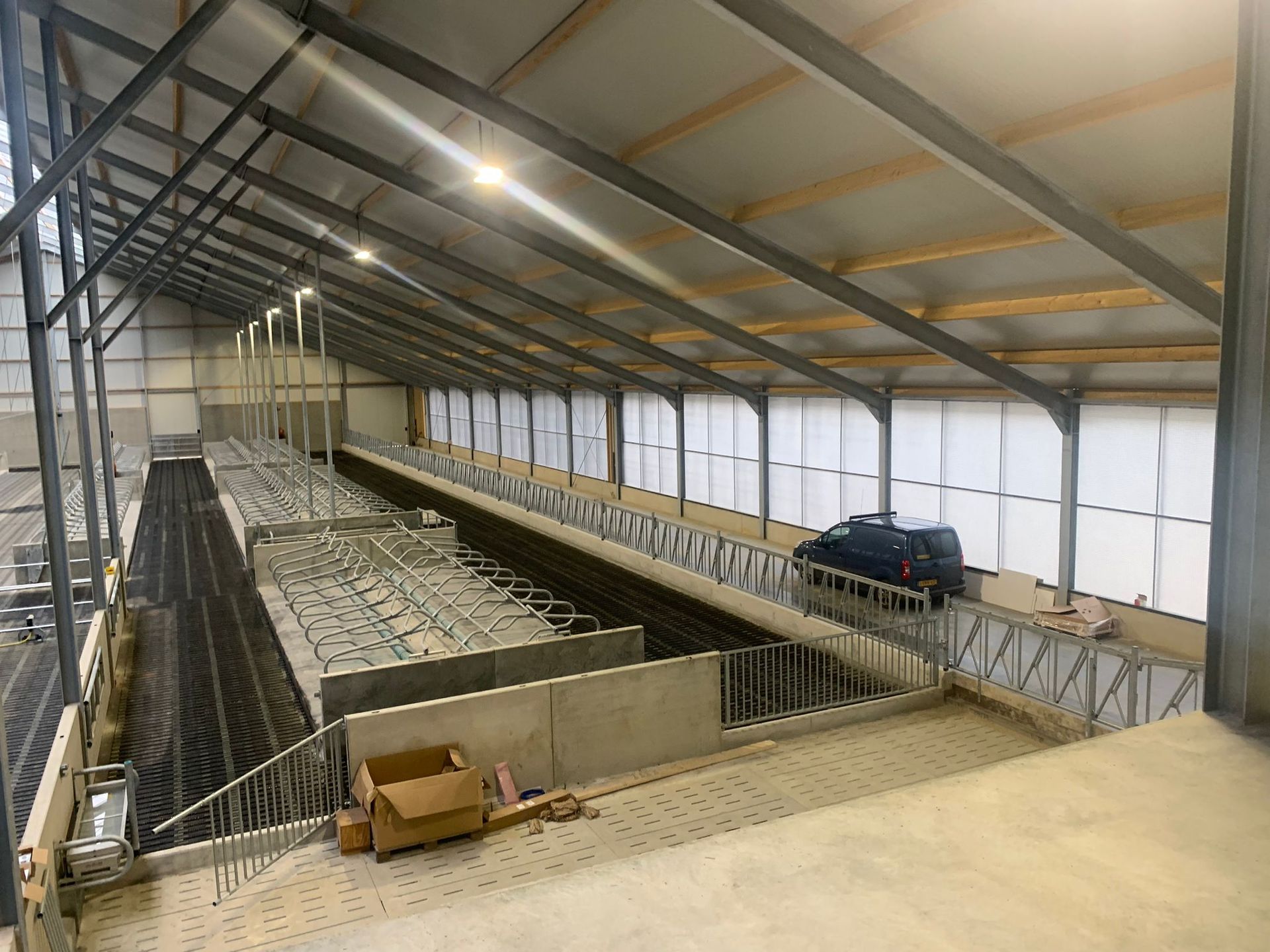 Een verhoogd uitzicht in een moderne, lege veestal met metalen balken, boxen en een donker voertuig dat erin geparkeerd staat.